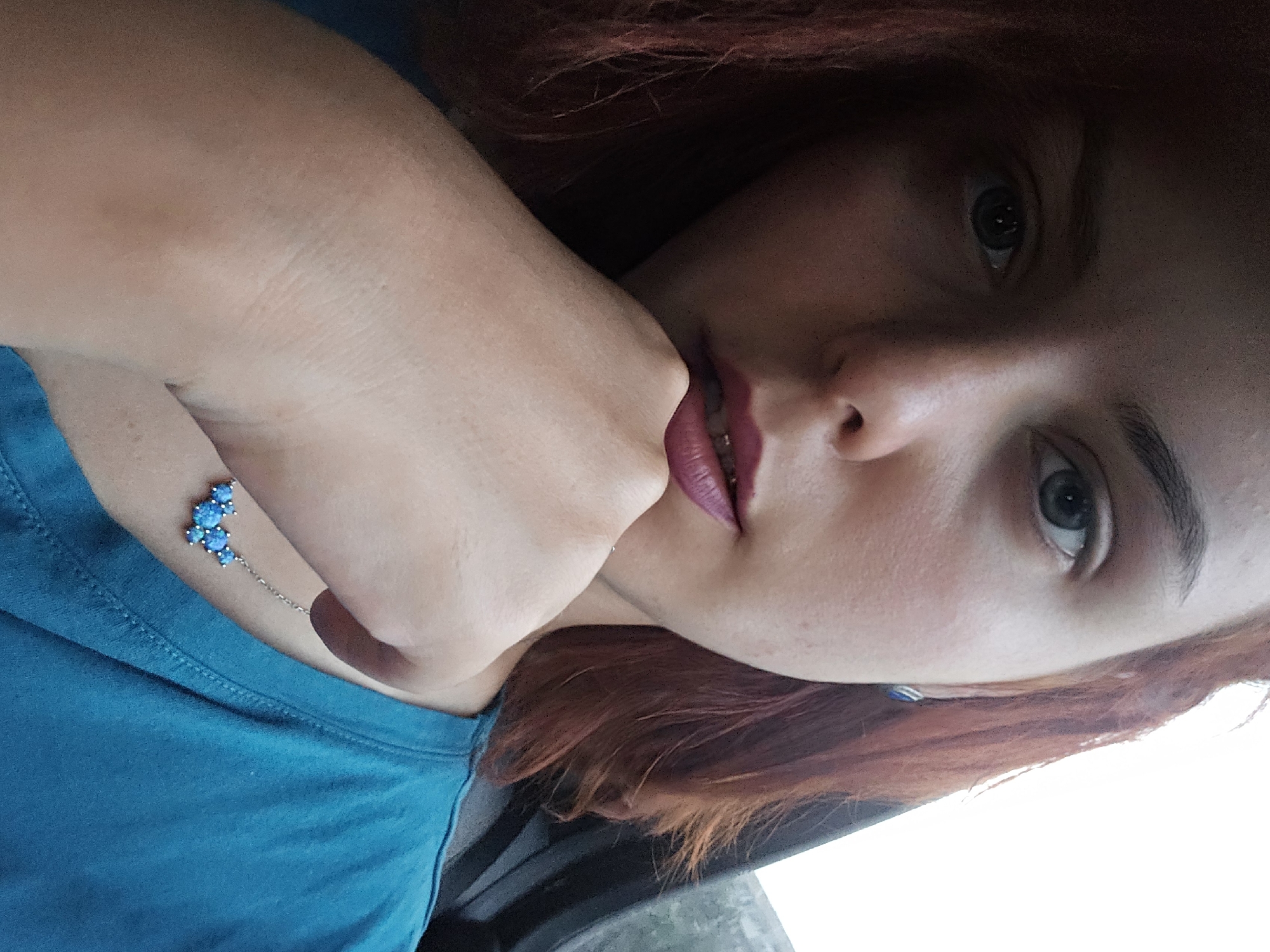 A young woman with auburn hair and blue eyes is captured in a close-up, low-angle selfie. She is wearing a teal t-shirt and a delicate silver necklace with blue stones. Her right hand is resting on her chin, and her lips are painted a deep berry color. The background appears to be the interior of a vehicle, with a dark window frame visible to the right and a bright, possibly overcast sky in the upper right corner. The lighting is soft, casting subtle shadows on her face.