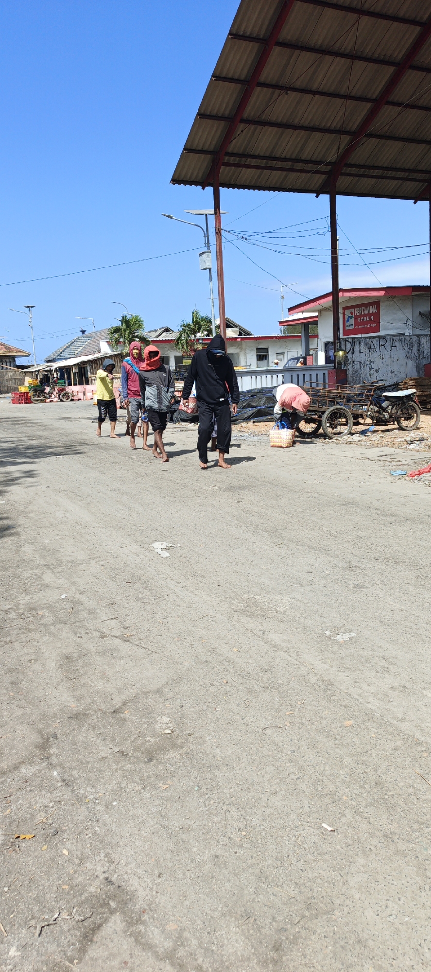In Puger, Indonesia, a group of four barefoot males walks along a dirt road under a clear blue sky. The man in the foreground wears a black hooded sweatshirt and black pants, with his face obscured by the hood. To his left, another man in a red head covering and a gray and black jacket walks with a group of two other men. One of these men wears a red head covering and a blue shirt, while the other is in a yellow shirt and black shorts. Behind them, a woman in a pink shirt bends over a wooden cart with two wheels, near a parked motorcycle and several large black tarps. Further in the background, buildings with red-trimmed roofs line the street, and one building displays a "PERTAMINA" sign. The road is a mix of dirt and gravel, with scattered debris.