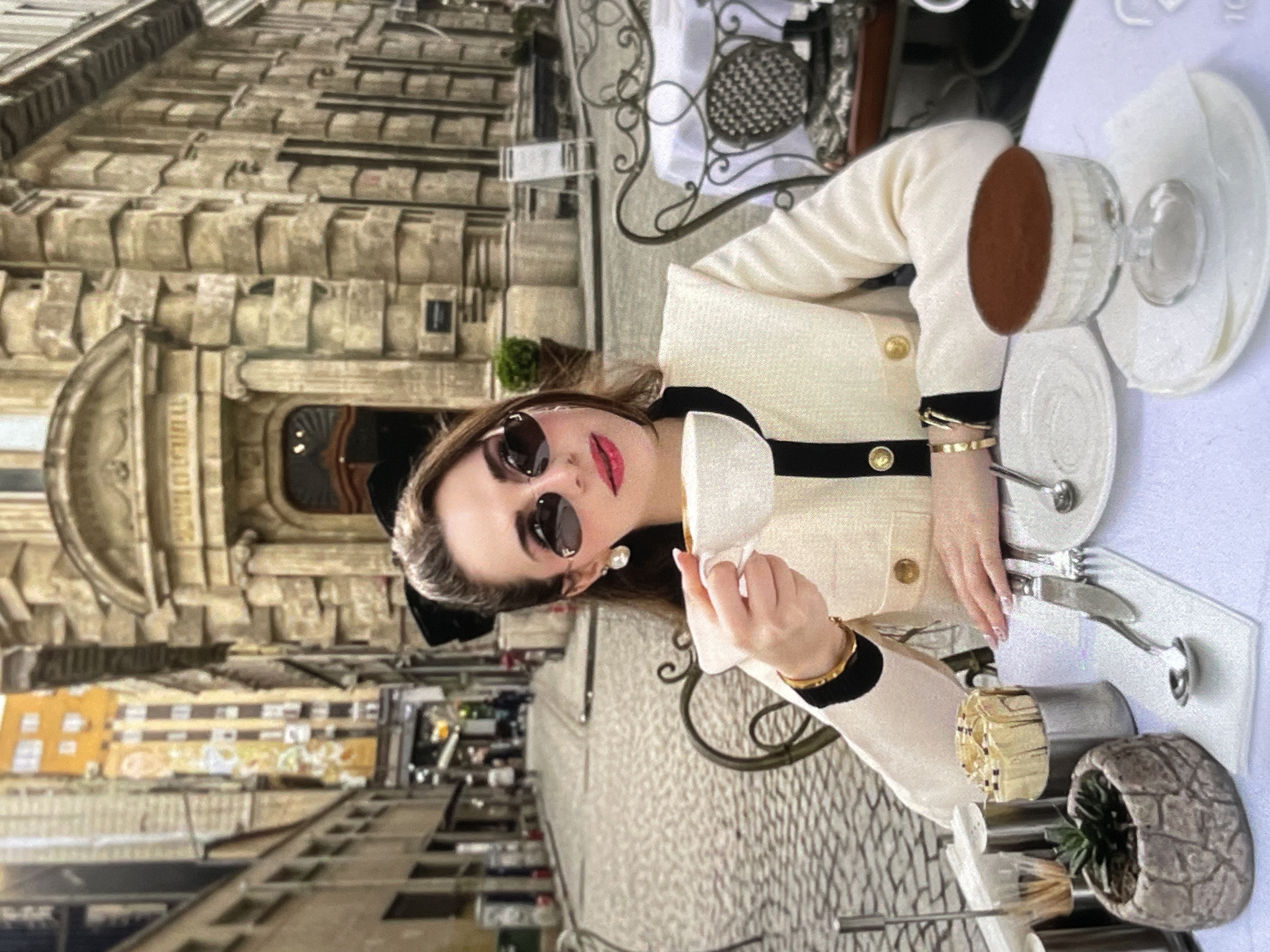 In an outdoor cafe setting on a cobblestone street in an urban environment, a woman with brown hair styled in a high ponytail, adorned with a black bow, sits at a table covered with a white tablecloth. She is wearing large, heart-shaped sunglasses with a dark frame, pearl earrings, and a cream-colored jacket with black trim and gold buttons. The woman holds a white teacup in her right hand, bringing it towards her lips. On the table, a small succulent plant in a stone-like pot, a container of toothpicks, and a dessert in a glass dish topped with cocoa powder are visible. The background reveals the architecture of old buildings lining the street, with a few blurred figures in the distance.