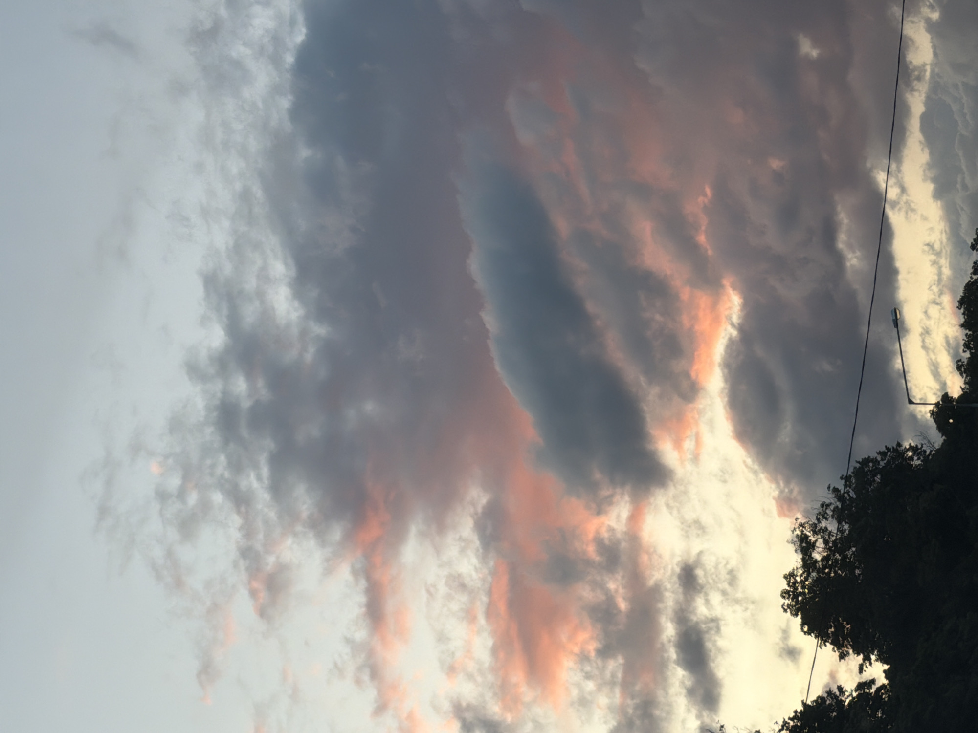 This is a vertical, medium shot of a twilight sky filled with dramatic clouds. The clouds are a mix of dark gray, soft pink, and pale yellow, with some areas of bright pink suggesting the setting sun. The sky transitions from a pale blue at the top to a warmer yellow-orange near the horizon. Silhouetted against the sky at the bottom are the dark shapes of trees and a portion of what appears to be a street light and electrical wires. The image was taken in Samara, Russia.