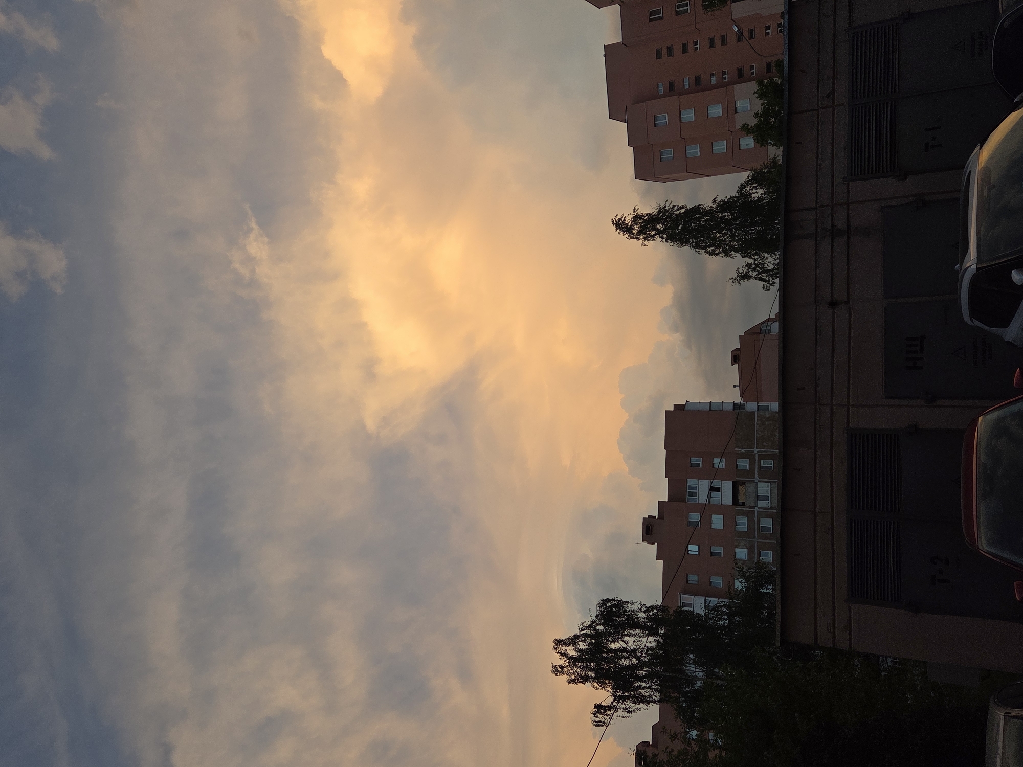 The image depicts a cityscape at dusk, with a sky filled with clouds illuminated by a warm, orange light, suggestive of sunset. On the right side of the frame, there is a building, appearing to be a residential structure, with multiple windows visible. Adjacent to the building is a row of garage units, their dark doors in stark contrast to the sky. In the foreground, parts of parked cars are visible, hinting at a residential area. A tree line partially obscures the building and the garage units, adding a natural element to the urban scene. The buildings and garages appear to be in an area of Unknown, Unknown.