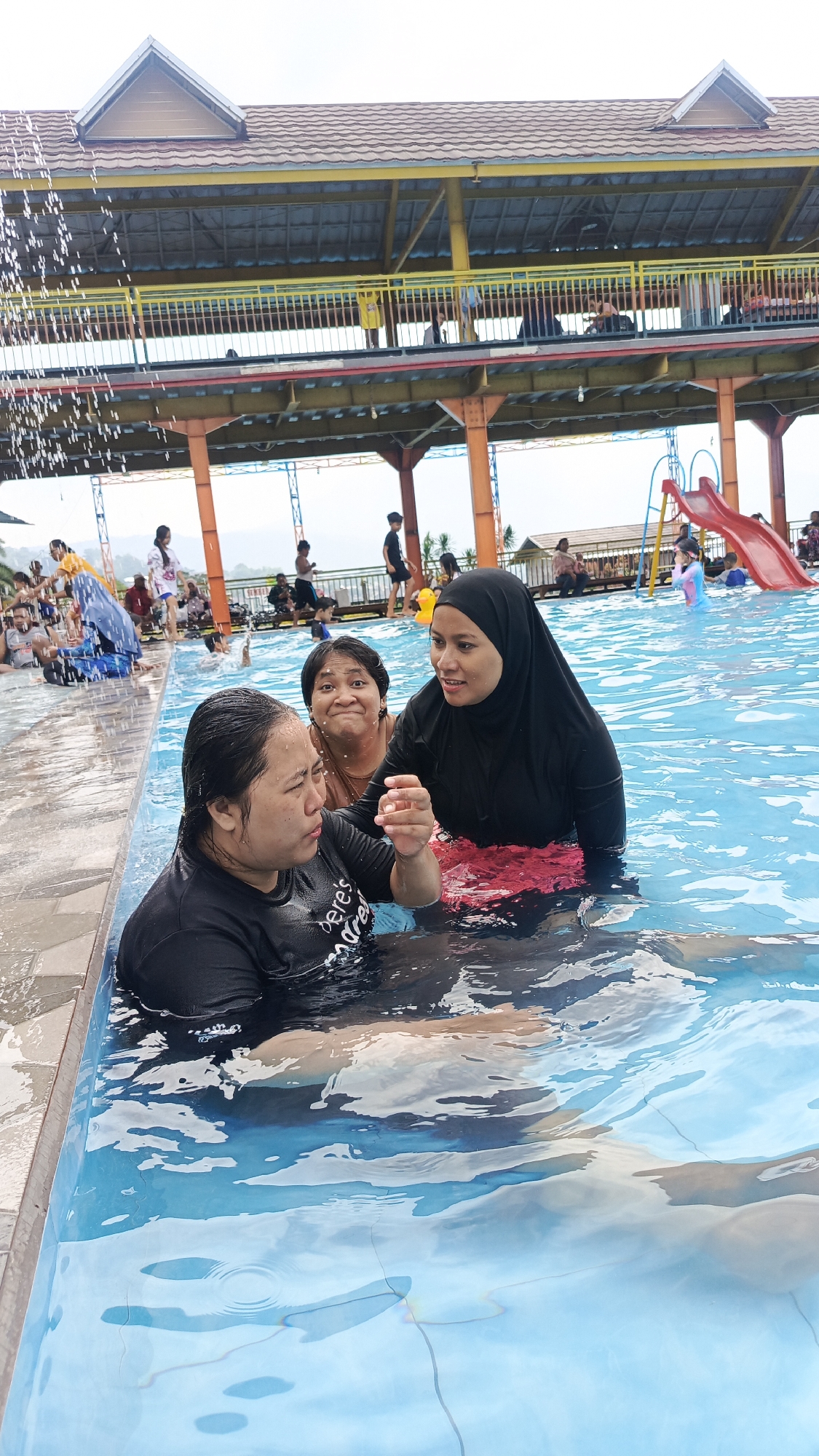 Three women are in a swimming pool. The woman on the right wears a black hijab and a red patterned skirt or bottom. The woman in the center has dark hair and is smiling at the woman on the right. The woman on the left is wearing a black t-shirt and is looking at the woman on the right. They appear to be in conversation.
The pool is located at Finna Golf and Country Club, in Prigen, Indonesia. In the background, people are enjoying the water park. There is a red slide and a covered structure with a yellow railing above the pool. Water is pouring down into the pool. There are hills in the distance.
