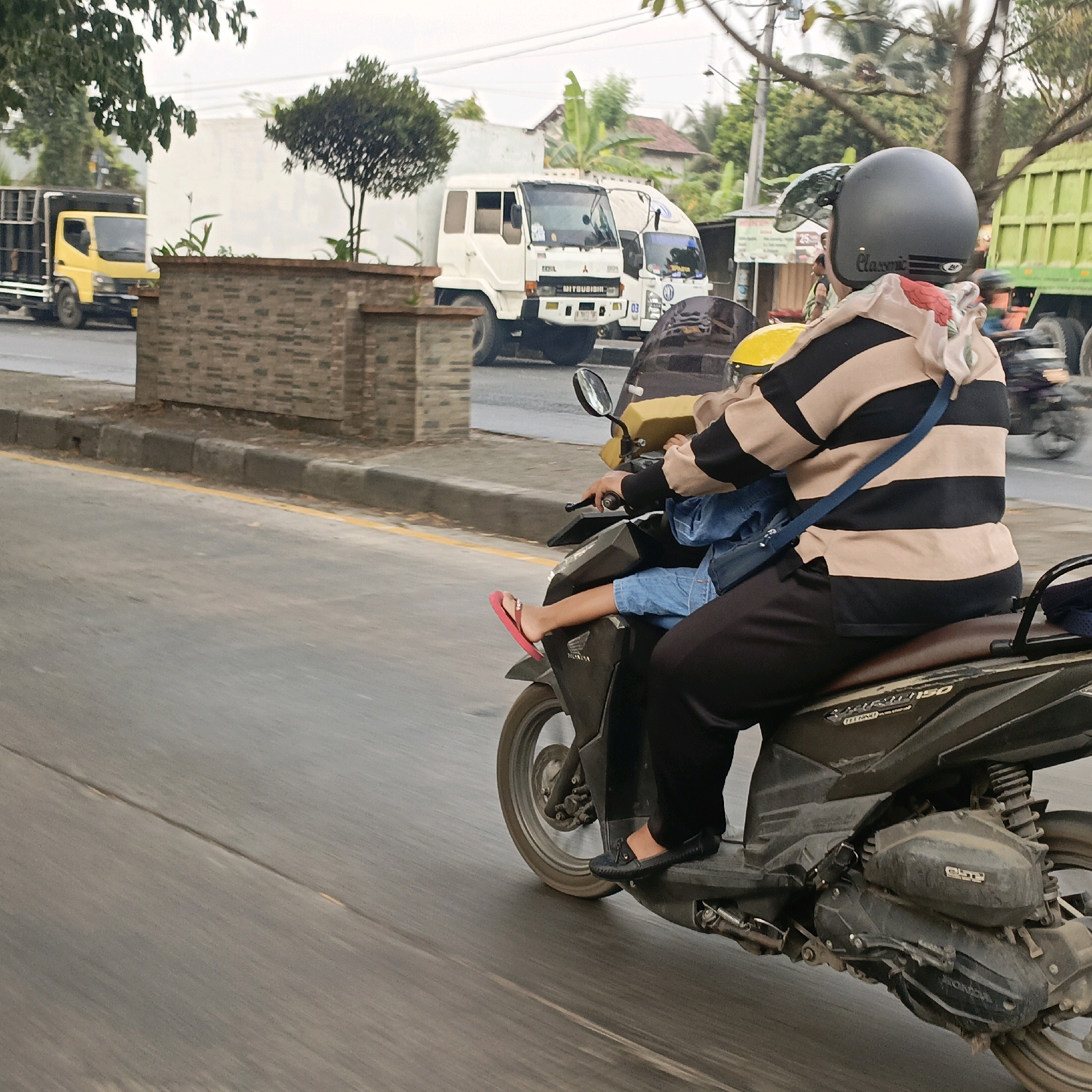 The image shows a motorcycle with two occupants riding on a street in Salatiga, Indonesia. The rider, wearing a black helmet with a silver visor and a black and beige horizontally striped long-sleeved shirt, is driving. A child, wearing a blue denim jacket and blue shorts, is seated in front of the rider and is wearing pink flip-flops. The child's right leg is extended, with the flip-flop visible. The motorcycle appears to be a Honda model. In the background, there are several vehicles, including a yellow truck, a white Mitsubishi truck, and a green truck. The environment suggests an urban or semi-urban setting with buildings and trees visible. The road surface is paved and shows signs of wear, with a yellow line marking the edge of the lane. The lighting suggests it is daytime, likely overcast or early morning/late afternoon.
