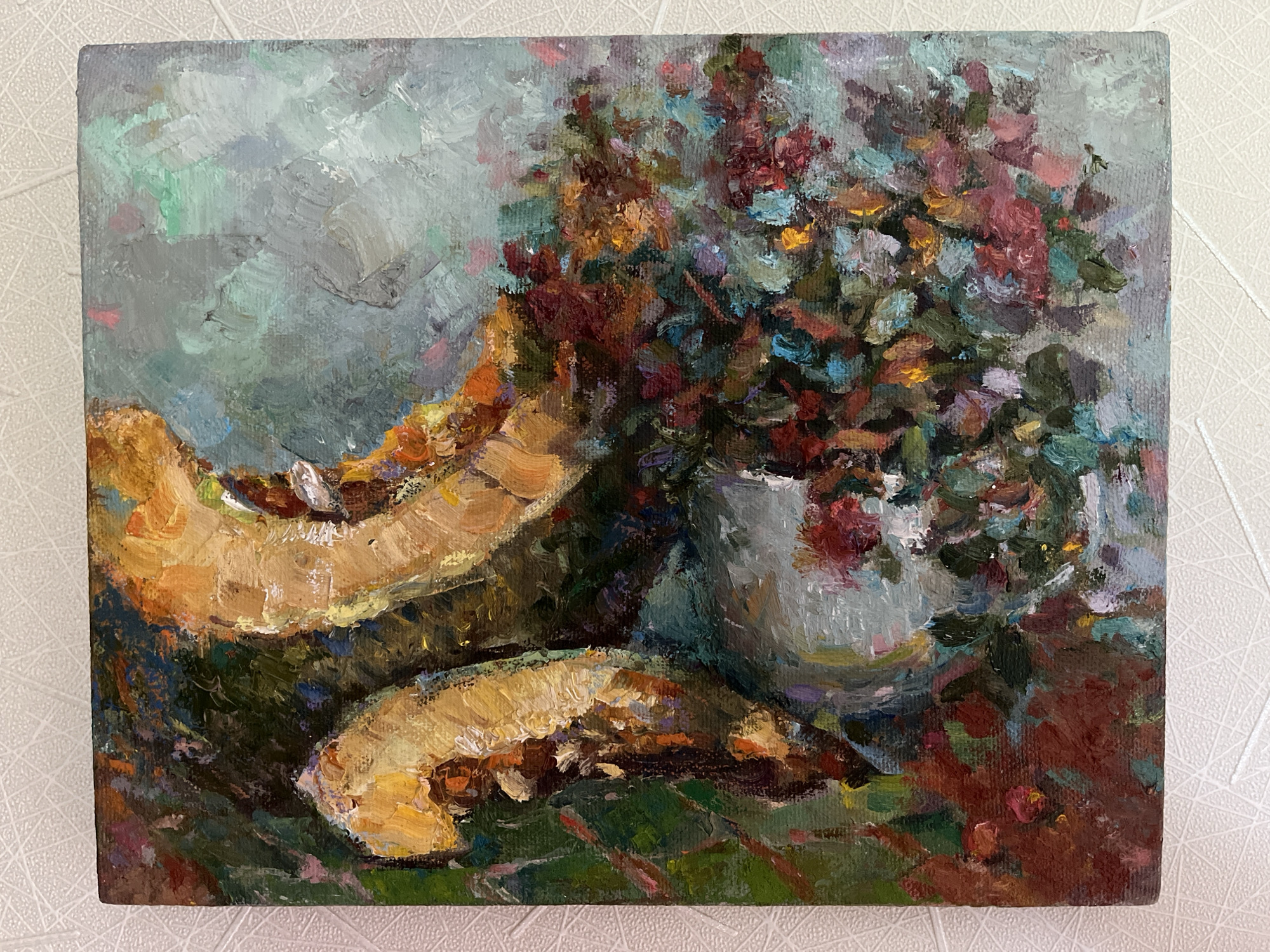 This is a square, impressionistic oil painting on canvas, featuring a still life arrangement. The primary subjects are two large, curved slices of a golden-yellow fruit, possibly pumpkin or squash, placed on a dark green surface. To the right of the fruit, a white vase overflows with a vibrant bouquet of flowers in various shades of red, burgundy, blue, and pink, with hints of yellow and orange. The background is a soft, impressionistic wash of muted blues, greens, and grays, suggesting a diffused light.
The painting captures a moment of domestic simplicity, evoking a feeling of warmth and abundance associated with harvest time. The thick, visible brushstrokes add texture and depth to the composition, giving the fruits and flowers a lively, almost tangible quality. The light appears soft and natural, possibly from an overcast day or diffused indoor lighting, with no strong shadows.
There is no visible text within the painting, nor any direct clues to a specific time of day or weather conditions beyond the gentle lighting. The setting is implied to be indoors, given the presence of the vase and bouquet. The overall mood is peaceful and slightly rustic, reflecting the painterly style and subject matter. The painting is presented on a white, textured surface, possibly a wall or tabletop.
