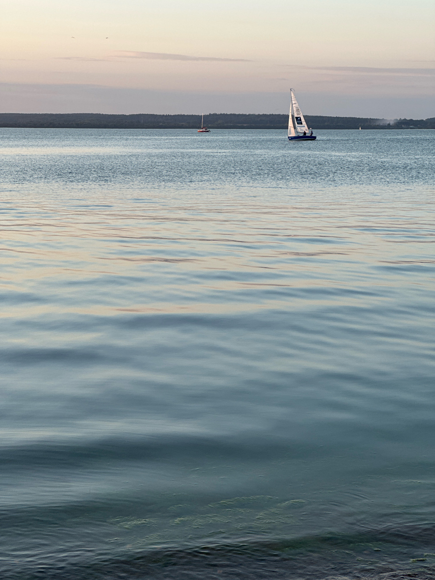 The image depicts a serene lake scene at dusk, likely located near Ждановіцкі сельскі Савет, Belarus. In the mid-ground, two sailboats are visible on the calm water. One sailboat, closer to the foreground and right of center, is white with blue accents and has two people on board. The other sailboat, further in the distance and to the left, is orange and white. The horizon is marked by a dark, tree-lined shoreline, with a muted, pastel-colored sky above. The foreground is dominated by the gentle ripples of the water, reflecting the soft light of the sky. The overall atmosphere is peaceful and tranquil.