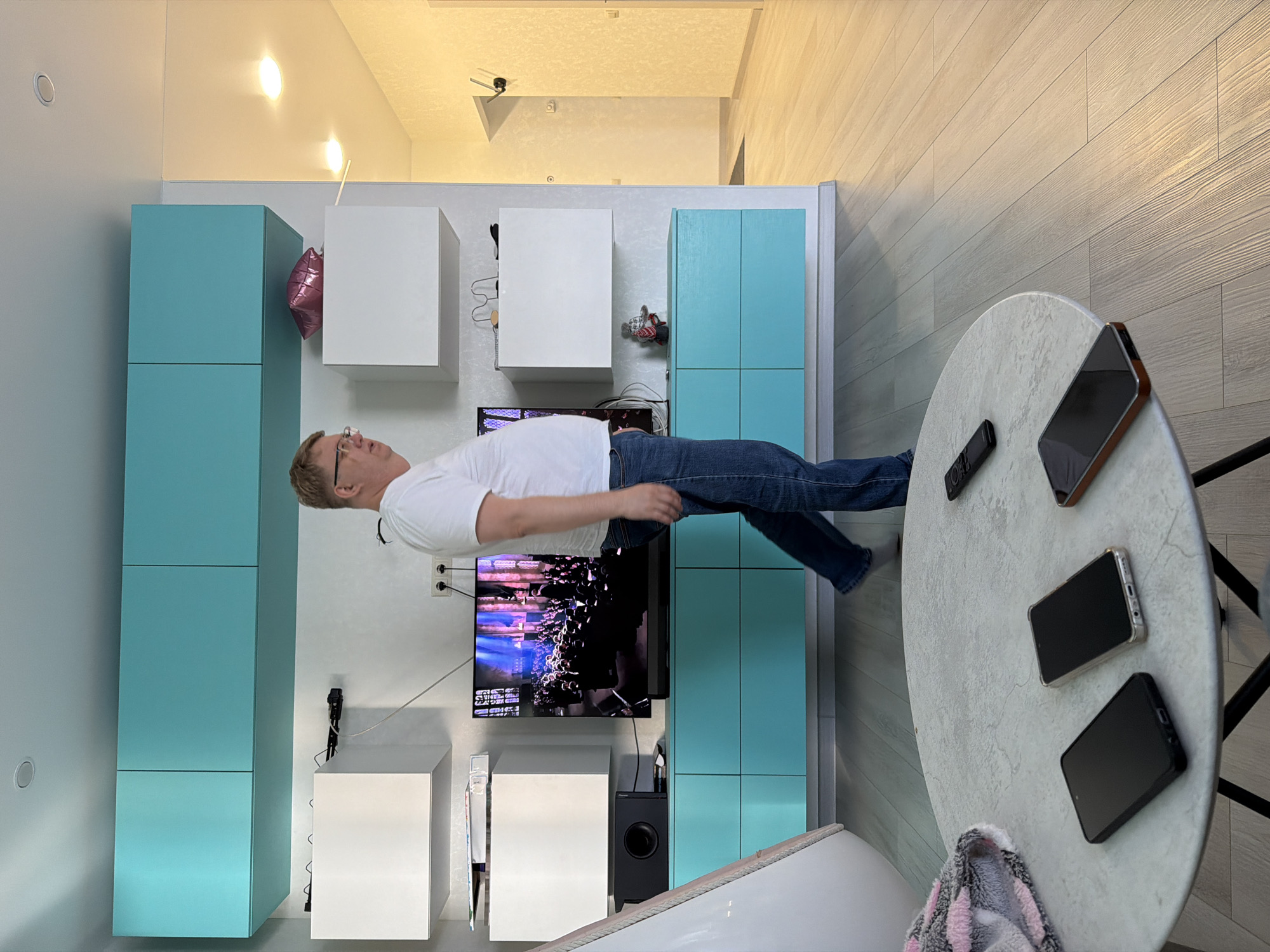 This media file depicts an indoor scene, likely a modern living room or apartment in Старое Михайловское, Russia. The central subject is a Caucasian man with light hair and glasses, wearing a white short-sleeved t-shirt and blue jeans. He is positioned horizontally across the frame, creating the illusion of floating in mid-air. This effect is a result of the photograph being rotated 90 degrees, suggesting the man was actually lying on his back on the floor when the image was taken, posing for the camera.
The room exhibits a contemporary aesthetic with light-colored walls and a floor covered in light-toned, wood-look laminate or tile planks. The ceiling features recessed lights and possibly track lighting. Along one wall, there are various storage solutions: several light turquoise (teal) cabinets are arranged vertically, interspersed with white cube-shaped shelving units. A flat-screen television is mounted on this wall, displaying what appears to be abstract or glitched digital art. Beneath the television is a black speaker or subwoofer, on which the brand "Pioneer" is visible.
In the foreground, to the right, stands a round, light-colored table. It holds a black remote control, at least three smartphones with dark screens, and a grey and pink striped fluffy towel or blanket. A pink foil balloon, possibly heart-shaped, is tucked between a teal cabinet and a white shelf unit. The scene is well-lit by artificial lights. The man's expression is neutral, contributing to the posed and artful nature of the image.