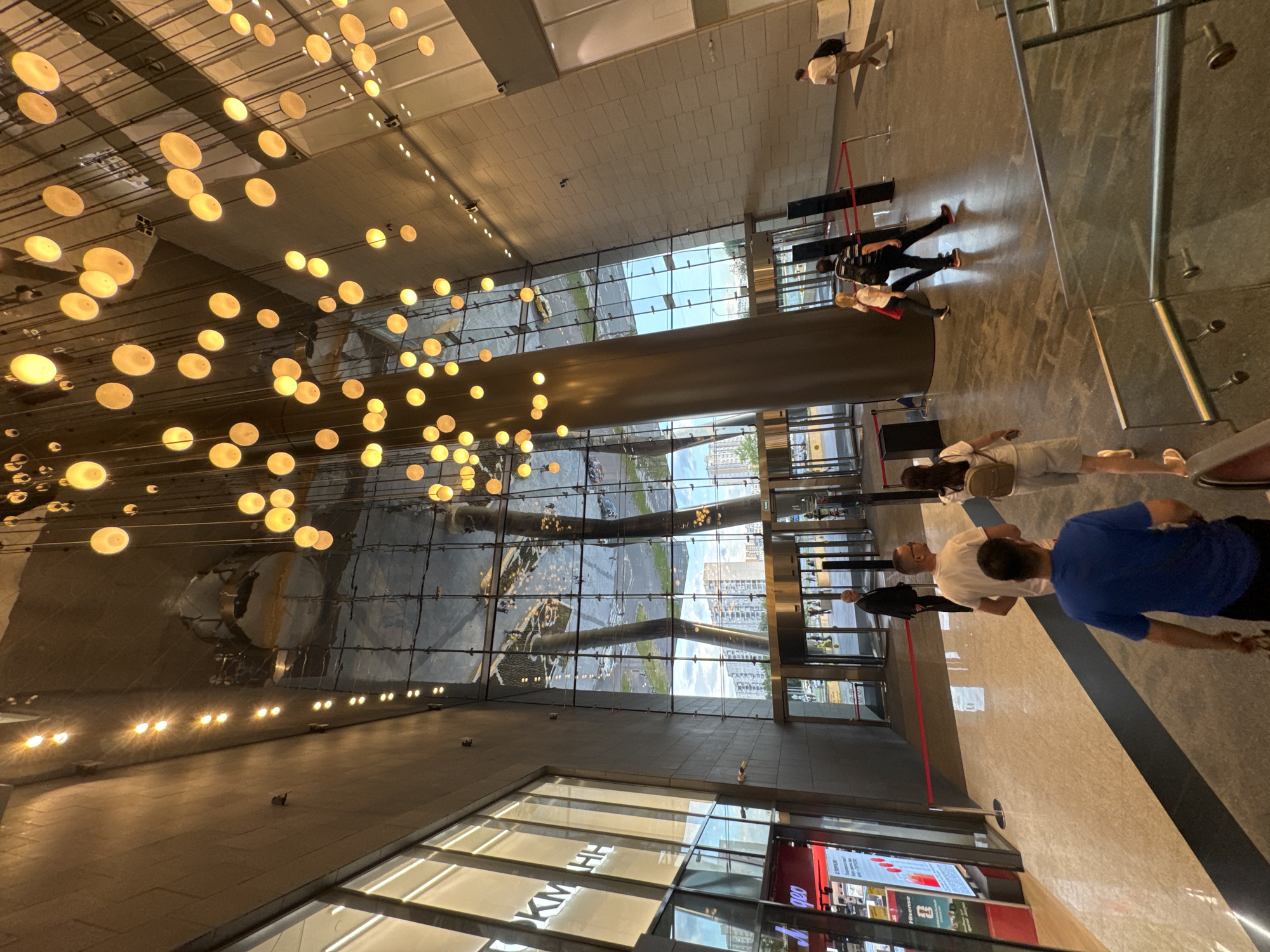 The image shows the interior of a modern building, likely a shopping mall or office complex, in Moscow, Russia. A large, open atrium features a high, reflective ceiling adorned with numerous pendant lights, creating a warm, ambient glow. The walls are clad in a light-colored, textured material, and large glass panels offer views of the exterior, where modern apartment buildings and a clear sky are visible. Several people are present, walking through the space, with one group in the foreground descending a staircase. The floor is a polished, marbled material with a dark stripe running through it. Red velvet ropes on stanchions guide pedestrian traffic. The overall impression is one of contemporary architecture and public space.