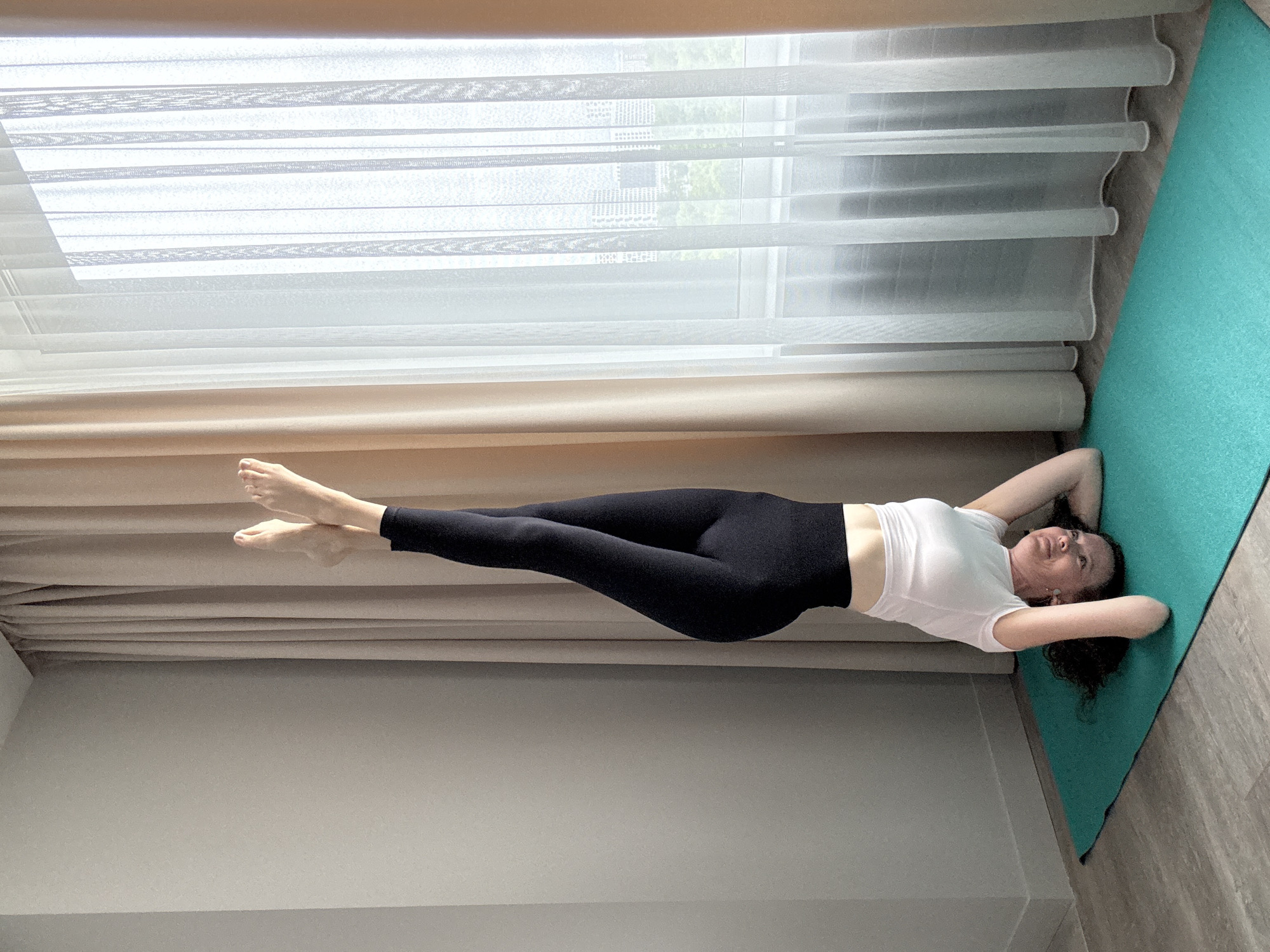 A woman is performing a headstand on a teal yoga mat in a room with beige curtains and a sheer white curtain covering a window. The woman has dark curly hair and is wearing a white t-shirt and black leggings. She is smiling and looking towards the camera. Outside the window, a cityscape is visible.