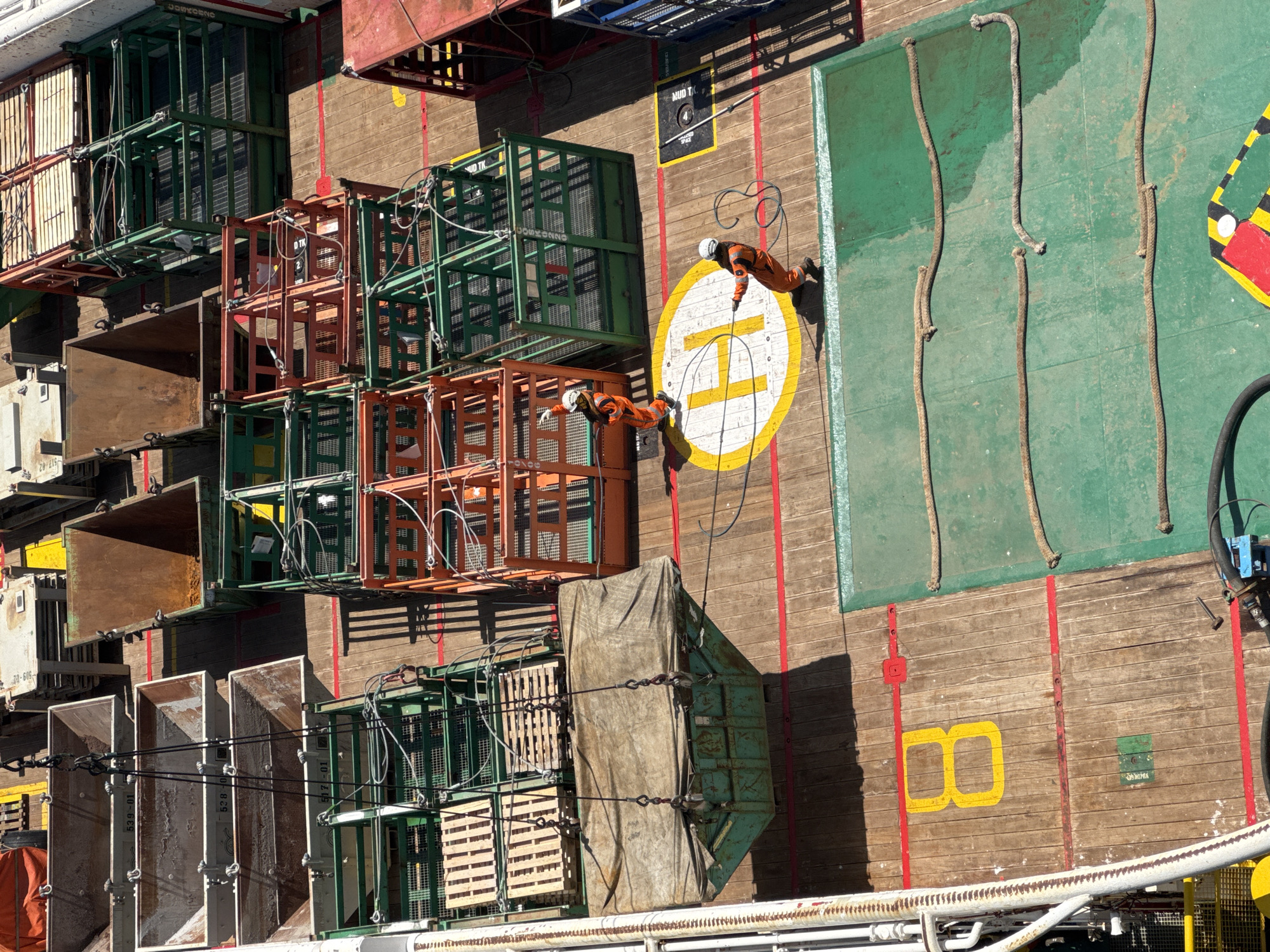 A high-angle medium shot captures a section of a ship's deck, cluttered with various cargo containers and industrial equipment, bathed in bright sunlight. Two individuals in orange protective suits and white hard hats are visible, engaged in tasks on the deck. One person, positioned on the left, is facing away from the camera, while the other, on the right, is facing left and appears to be operating a hose. The deck itself is a mixture of wooden planks and green painted surfaces, marked with yellow and red lines, along with stenciled numbers and symbols. Notably, a large yellow "H" is painted on the deck, indicating a helipad. Several green and orange metal cages, some containing wooden pallets, are stacked on the deck, suggesting active cargo handling or storage. A large, tarp-covered object is suspended by cables, adding to the dynamic nature of the scene. The overall impression is one of industrial activity on a maritime vessel. The scene does not provide any specific information or visual cues that would indicate its location within the city of Unknown, Italy.