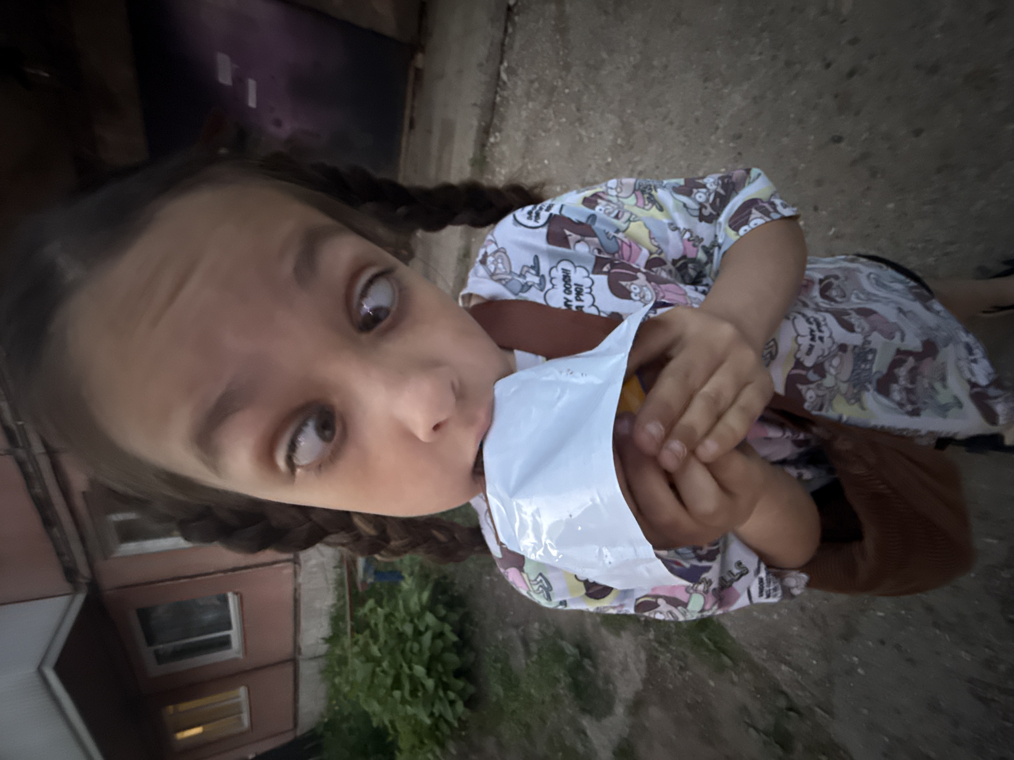 A young girl with brown braided hair is eating something from a white plastic wrapper. She wears a patterned white shirt. The background includes a pink building with windows and green foliage. The image appears to be taken outdoors. I cannot confirm the location as Votkinsk, Russia, without additional information or context.