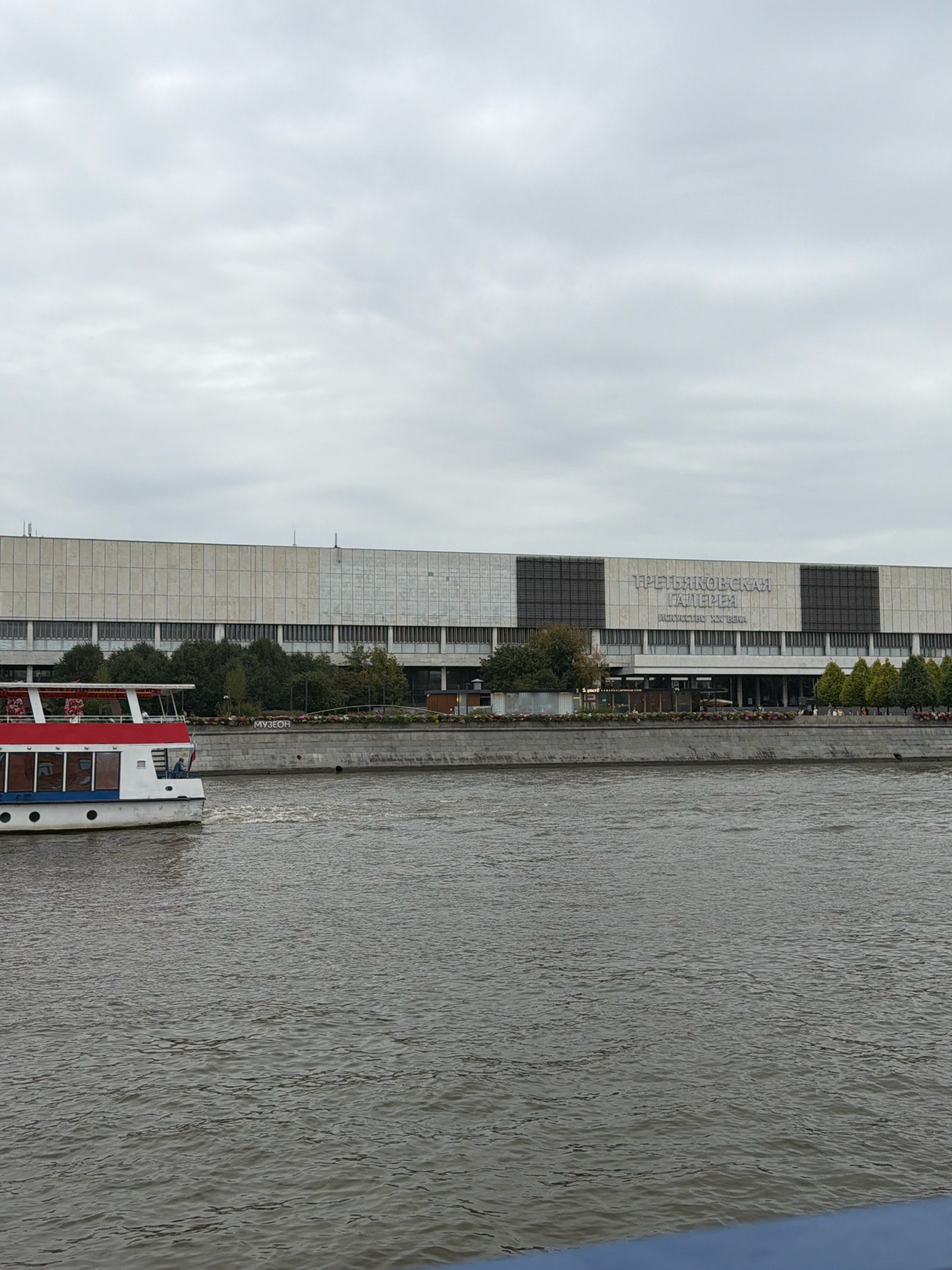 This is a photograph taken from a body of water, likely a river, looking towards a large, modern building in Moscow, Russia. The building has a long, horizontally-oriented facade constructed of light-colored stone or concrete panels. Large windows are visible along the lower stories, and a section of the facade in the center is darker, possibly indicating a different material or architectural feature. The building has signage with Russian text, the words "ТРЕТЬЯКОВСКАЯ ГАЛЕРЕЯ" (Tretyakov Gallery) are prominently displayed in large, light-colored lettering, with smaller text below reading "ИСКУССТВО XX ВЕКА" (Art of the XX Century).
In the foreground, on the left side of the image, a white and red tour boat is moving away from the viewer and towards the right. The boat has multiple decks with windows and decorative elements, including flowers on the upper deck. The water is a murky brown-gray color with ripples and waves.
Along the waterfront in front of the building, there is a stone embankment with trees and some flowering plants. People are visible walking along the embankment and near the entrance to the building. The sky is overcast with gray clouds, suggesting a cloudy day. The overall scene depicts a view of the Moscow River with a prominent cultural institution.
