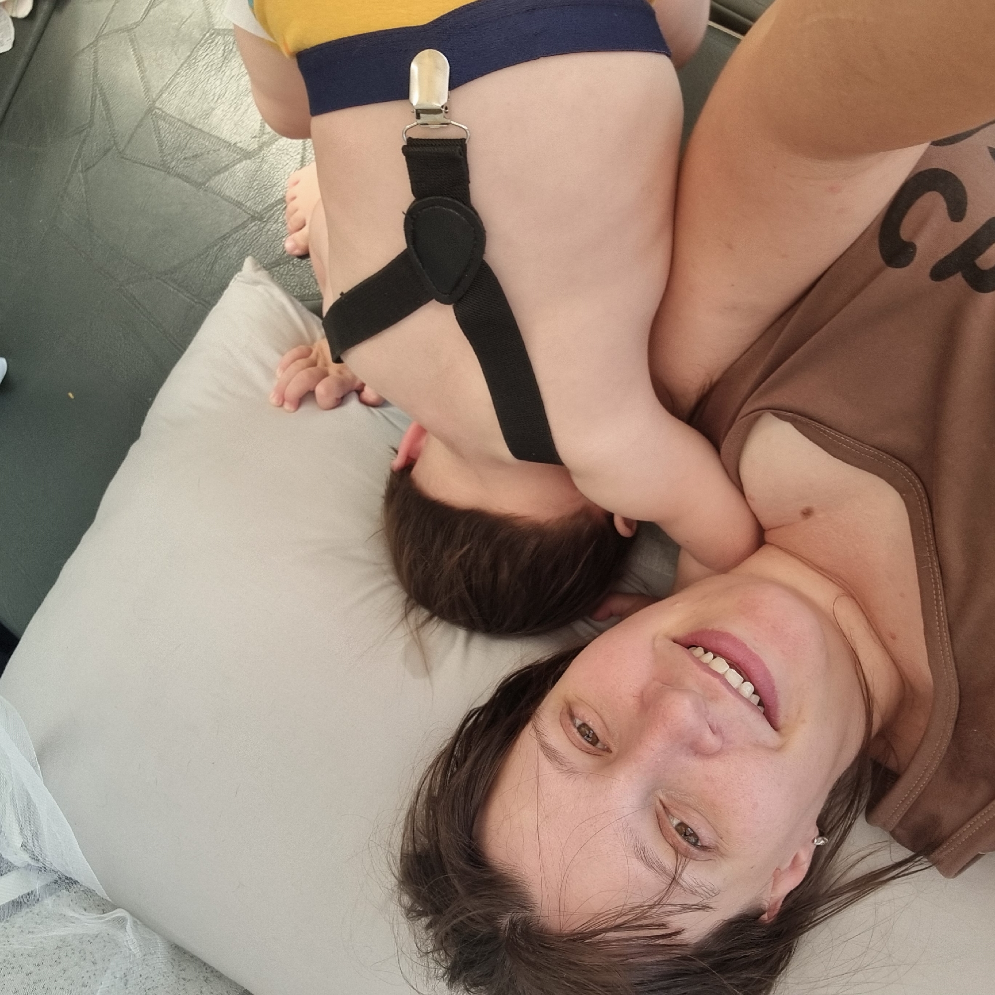 In Dunnae-myeon, South Korea, a woman is lying on a light gray pillow with her head propped up. She is smiling and looking at the camera. To her right, a young child is sleeping on the pillow, with their head resting on the woman's chest. The child is wearing yellow shorts with a blue waistband and black suspenders. The woman is wearing a brown tank top. The background appears to be a dark surface, possibly a couch or a bed.