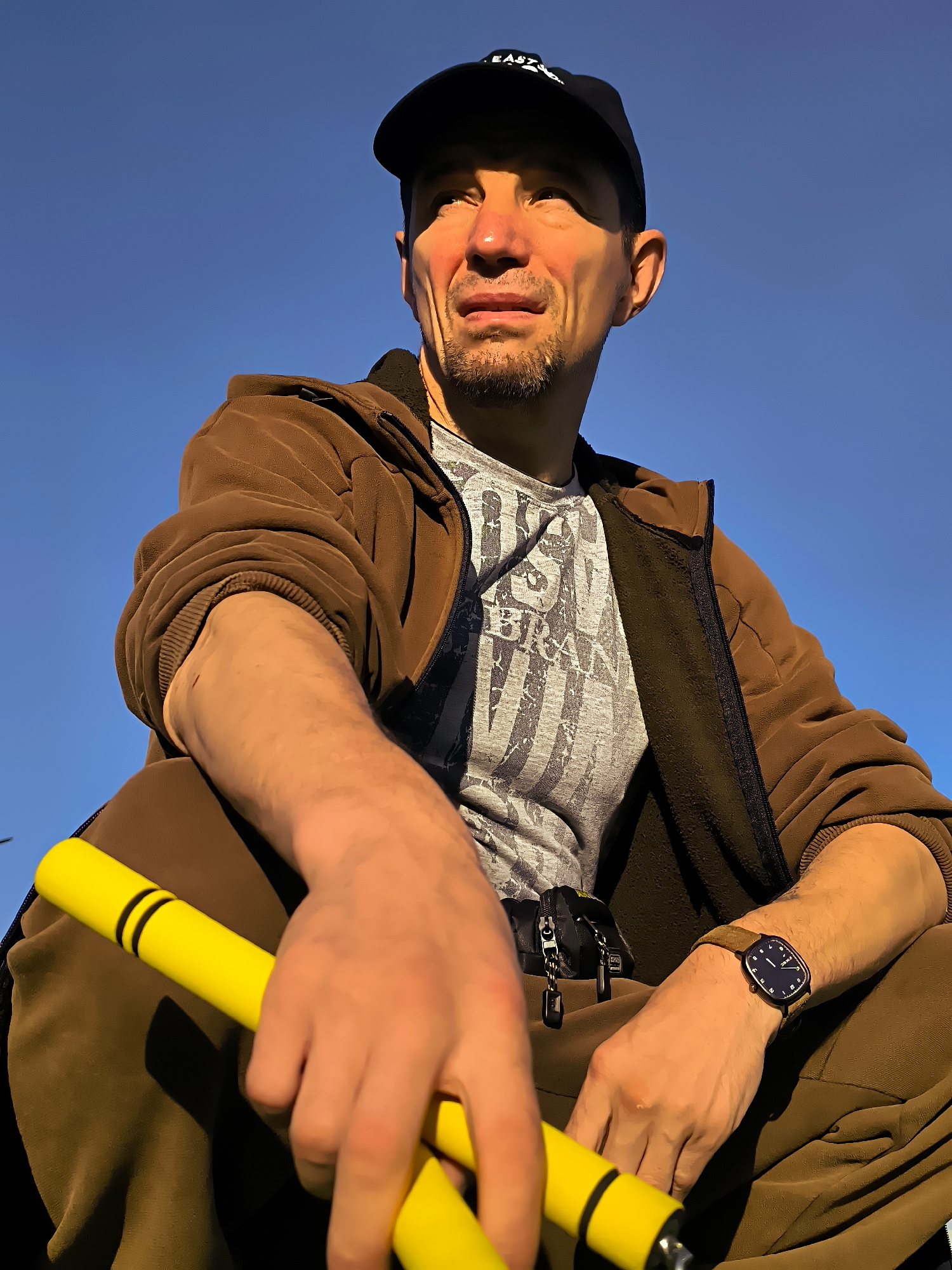 A medium shot from a low angle captures a Caucasian man sitting outdoors against a clear blue sky. He wears a dark baseball cap, a brown zip-up jacket over a grey t-shirt, and brown pants. A light-colored watch is visible on his left wrist. He holds a yellow object with black bands in his hands. A black bag with zippers is attached to his pants. The man's gaze is directed upwards and to the left, and his expression is thoughtful. The lighting suggests it is daytime. The image does not provide any specific details about the city of Qarağandı city administration, Kazakhstan.