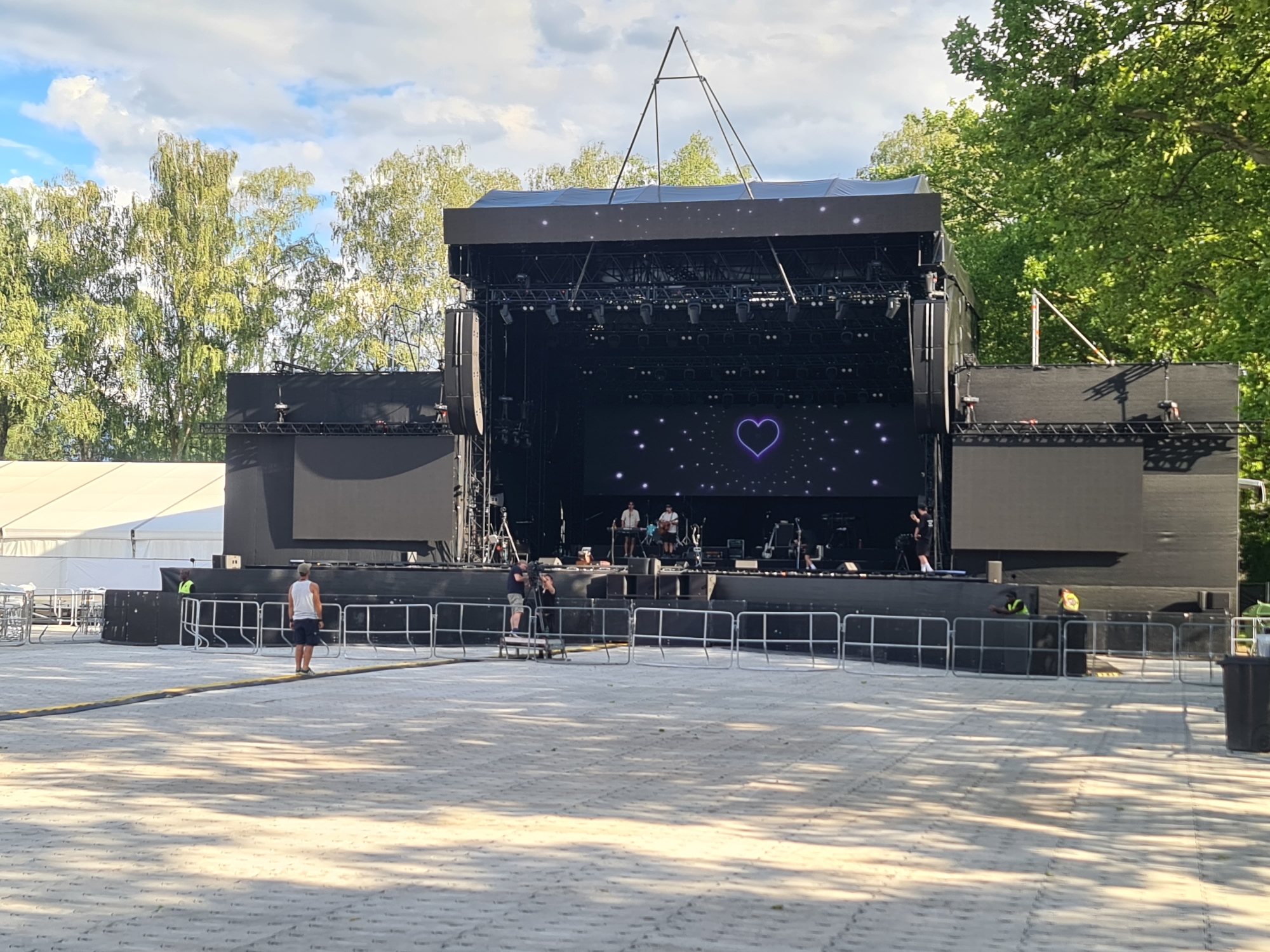 The image depicts an outdoor concert stage setup in Klagenfurt, Austria. The stage is large and black, with a large LED screen displaying a purple heart icon amidst a scattering of white dots. Several speakers are mounted on the sides of the stage. In front of the stage, there is a metal barricade, and behind it, a few people are visible on the stage, appearing to be performers or technicians. The ground in front of the stage is paved with light-colored interlocking bricks. Trees with green foliage are visible in the background, and the sky is partly cloudy with blue patches. Two individuals are standing in the foreground, one wearing a white tank top and shorts, and the other, partially visible, appears to be wearing a yellow vest, possibly a security guard. A large white tent structure is also visible to the left of the stage. The scene appears to be set up for an event, but it is not currently active.