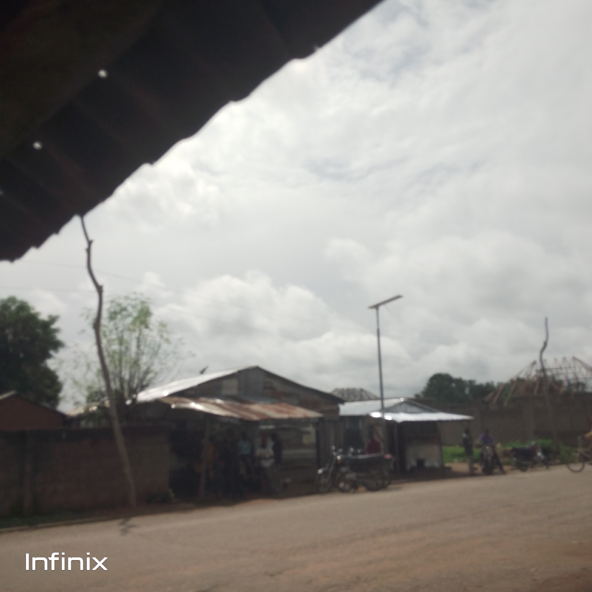 The image depicts a roadside scene in what appears to be a rural or semi-urban area. The sky is overcast with diffused light, suggesting it is daytime, possibly late morning or afternoon. In the foreground, a dirt or gravel road runs horizontally across the frame.
On the left side of the road, there are several structures that look like market stalls or small shops, some with corrugated metal roofs. People are visible near these structures, though their features are indistinct due to the image's blurriness. Two motorcycles are parked in front of one of the stalls.
Further down the road, on the right, a street light is visible, casting no discernible shadow. More structures, possibly houses or shops under construction, are present in the background, along with some trees. The overall setting suggests a commercial or residential area situated along a road.
The image is significantly out of focus, which makes it difficult to discern precise details of the people, objects, or the specific environment. The blurriness gives the scene a somewhat hazy or dreamlike quality.
The word "Infinix" is visible in the bottom left corner of the image, in white text. This likely indicates the brand of the device used to capture the photograph. The image does not convey any specific emotional tone due to the lack of sharp details.