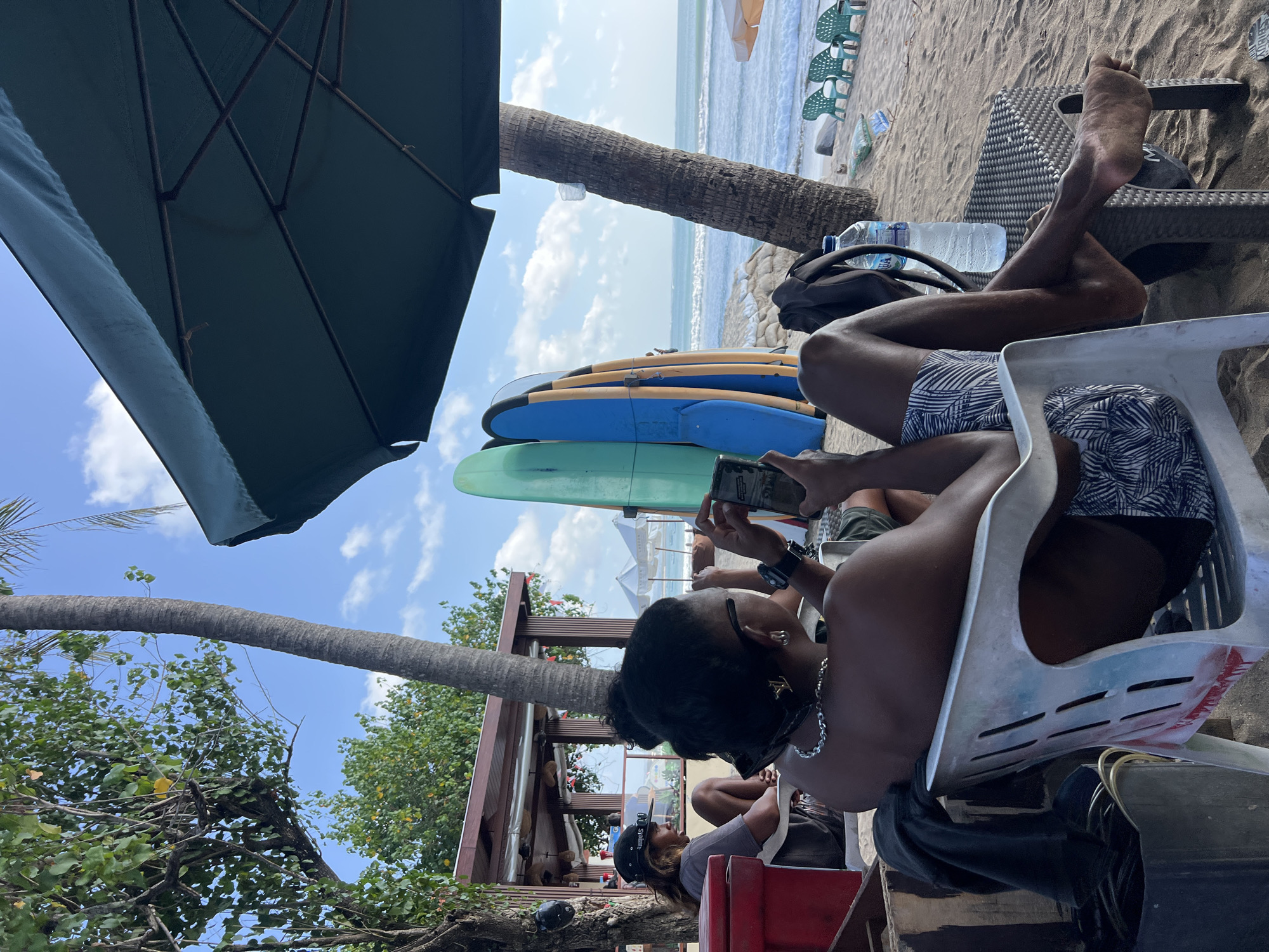 In Kuta, Indonesia, on a sandy beach, a person sits in a white plastic chair, looking at their phone. They are wearing patterned swim trunks and a silver chain necklace. Next to them, another person in a black hat and grey t-shirt sits in a red cooler. Several surfboards are propped up behind them. In the background, the ocean waves crash onto the shore, and palm trees sway under a blue sky with scattered clouds. The scene is casual, with beach chairs and umbrellas visible in the distance.