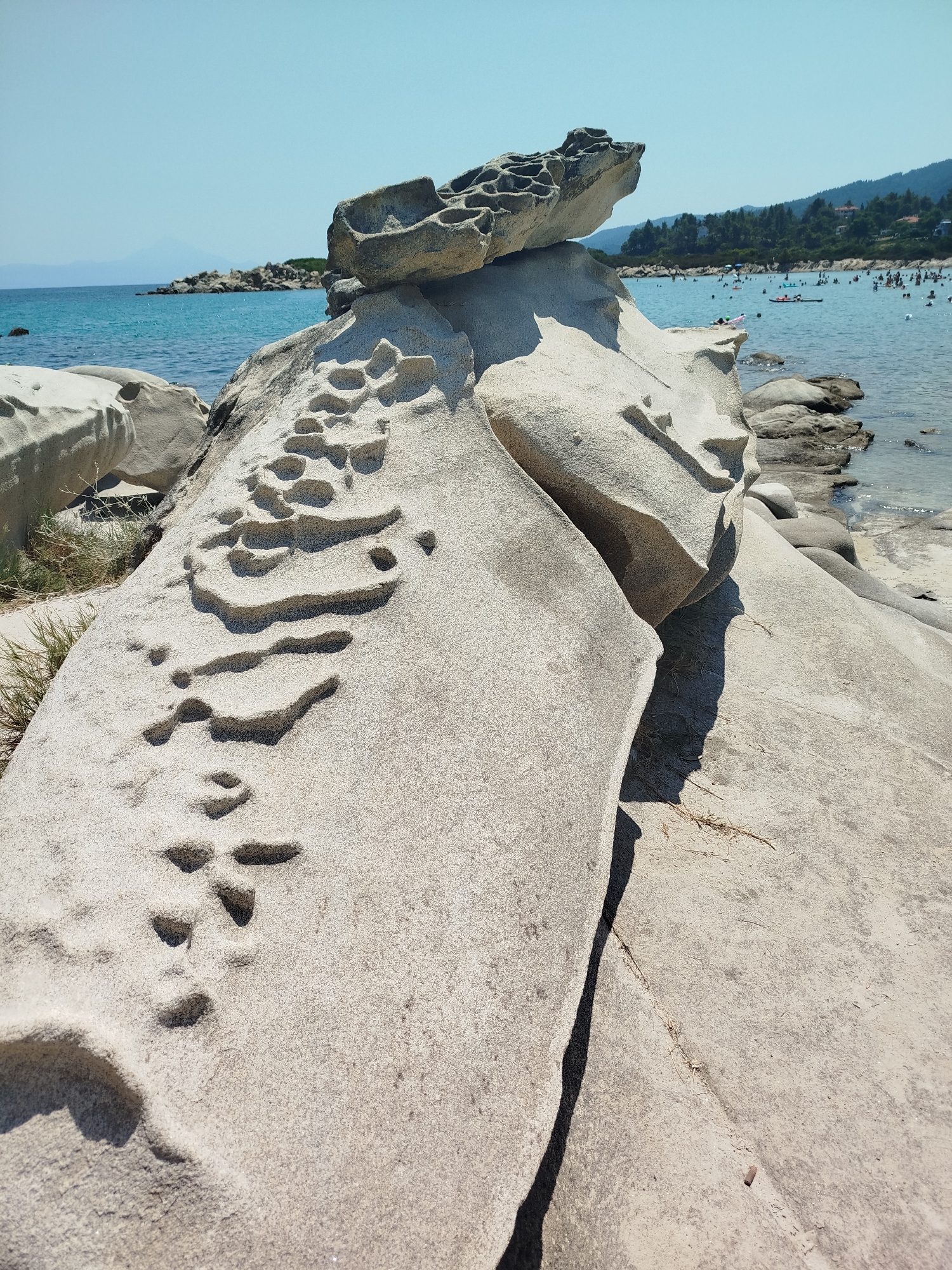 The image depicts a close-up view of unusual rock formations on a sandy beach, with the turquoise sea and a populated coastline in the background. The foreground is dominated by light-colored, weathered rocks that exhibit a honeycomb-like texture with numerous small, irregular holes. These rocks are sculpted by natural erosion, creating a unique, almost artistic, appearance. One large rock in the foreground has a flattened, layered top with more pronounced hollows, resembling a hollowed-out surface.
To the right of these prominent formations, the sea stretches out, appearing calm and clear. Further in the distance, the coastline is lined with lush green trees and buildings, suggesting a developed area. The water is populated with numerous people swimming, sunbathing on the shore, and engaging in water activities, including what appears to be a kayak or small boat. The sky is a clear, pale blue, indicative of a sunny day.
The specific location is Sithonia Municipal Unit, Greece. This area is known for its beautiful beaches and distinctive geological features, such as the ones captured in this photograph. The scene suggests a popular tourist destination where natural beauty coexists with human leisure activities.