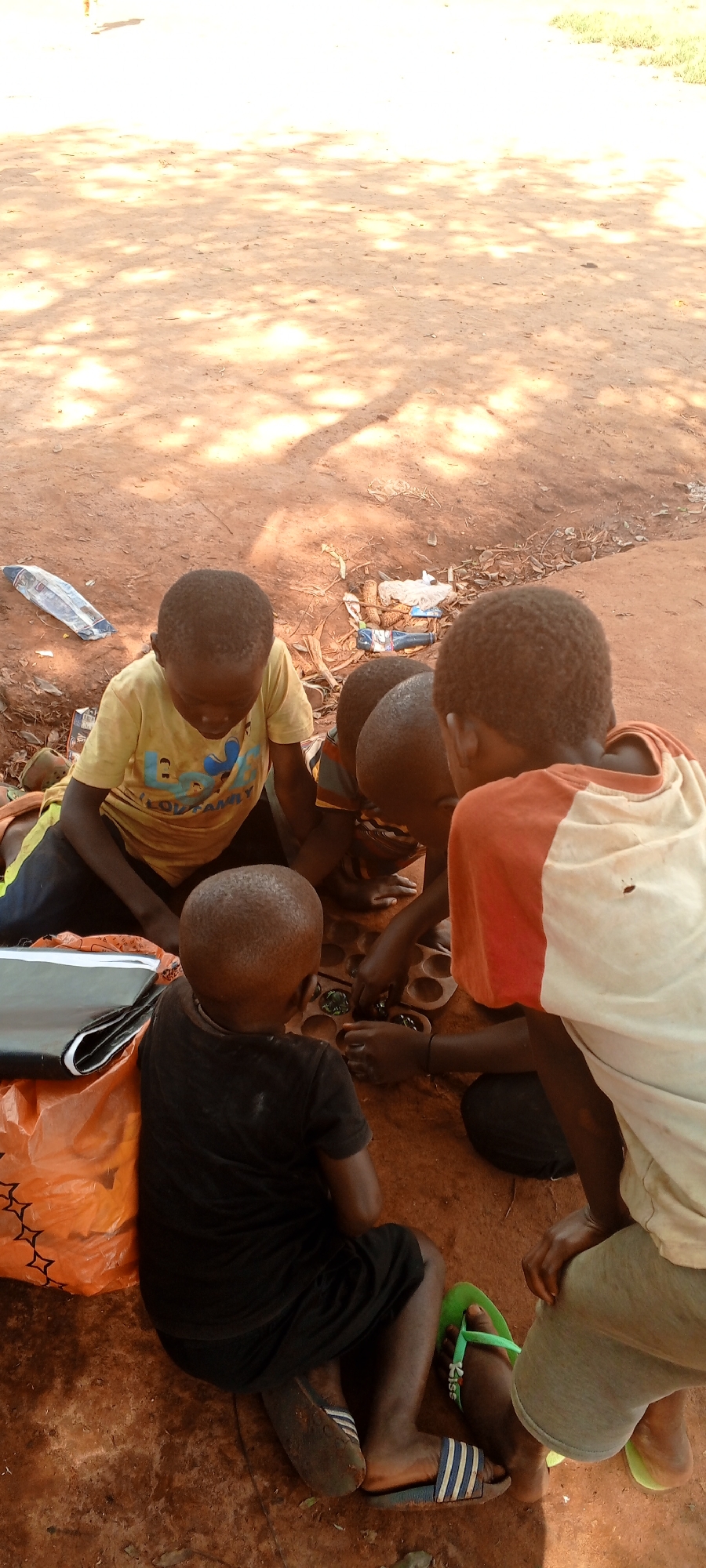 The image depicts five individuals, likely children or adolescents, gathered on a reddish-brown dirt ground in Ngalula, Democratic Republic of the Congo. Four individuals are actively engaged around a wooden board game, possibly a Mancala variant, with multiple carved depressions containing small green and dark playing pieces.
One child, wearing a yellow t-shirt with a blue graphic and black shorts, sits on the left, looking down at the game. Another child, seen from behind, wears a black t-shirt and dark shorts, also focused on the game. A third child, wearing an orange and white t-shirt, is positioned on the right, observing the game. Another partially visible child in a striped shirt is also present near the game board. Footwear includes blue and white striped slides and green flip-flops.
To the left, an orange plastic bag with a black object on top is visible. In the background, scattered on the dirt, are various pieces of litter including a clear plastic bag/bottle, white plastic remnants, dried leaves, and a small blue bottle. The ground is unpaved reddish-brown dirt, illuminated by dappled sunlight suggesting overhead foliage, with sparse green vegetation visible in the upper background.