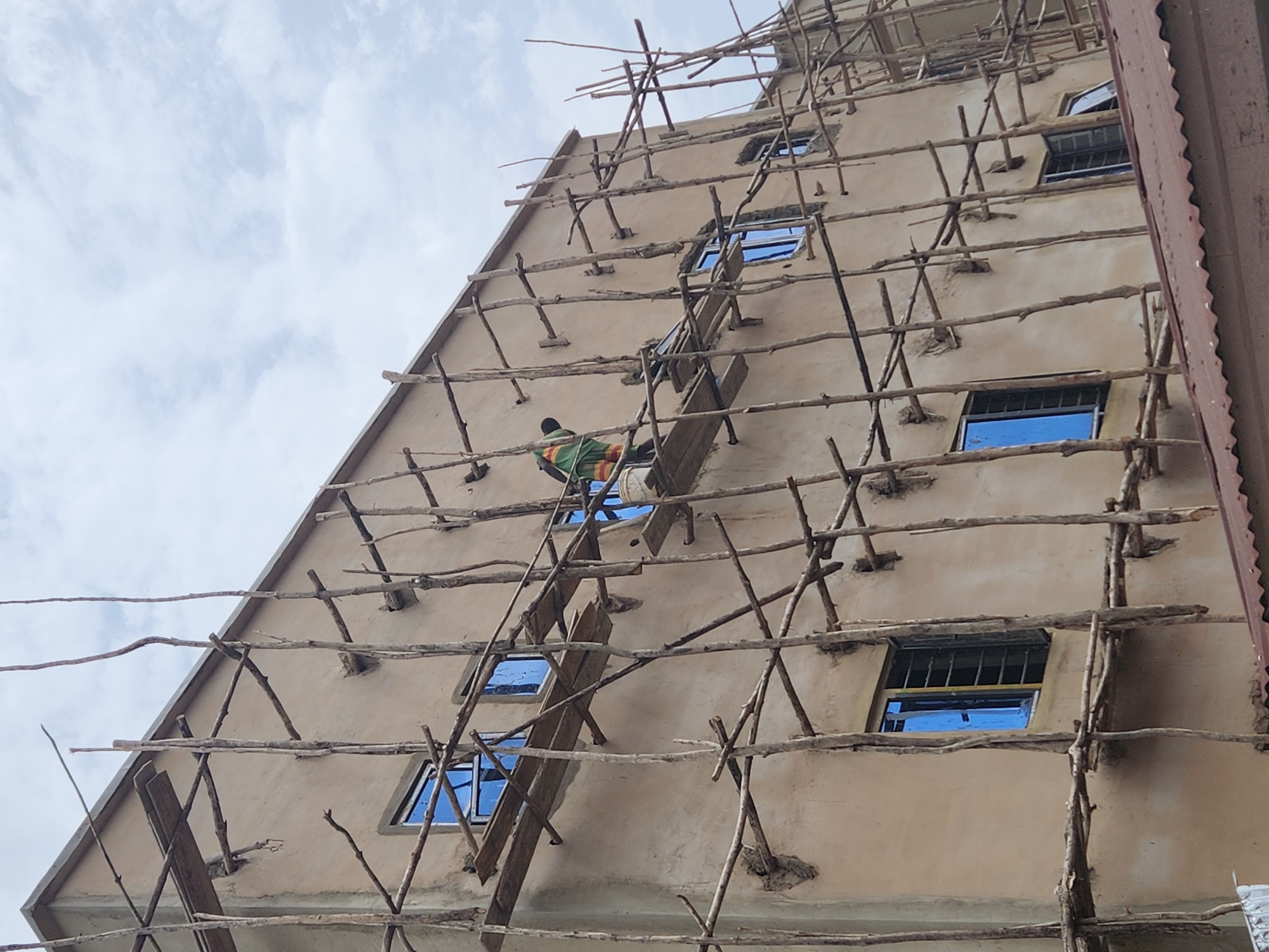 A low-angle, medium shot captures a building under construction in Jos, Nigeria. The exterior is light brown, with several windows reflecting the blue sky. A rudimentary scaffolding system, constructed from thin branches and sticks, is erected against the building's facade.
A man wearing green is situated on the scaffolding, possibly working on the building. The sky is visible in the background, with scattered clouds. The overall impression is one of a developing area, with construction practices utilizing locally available materials.