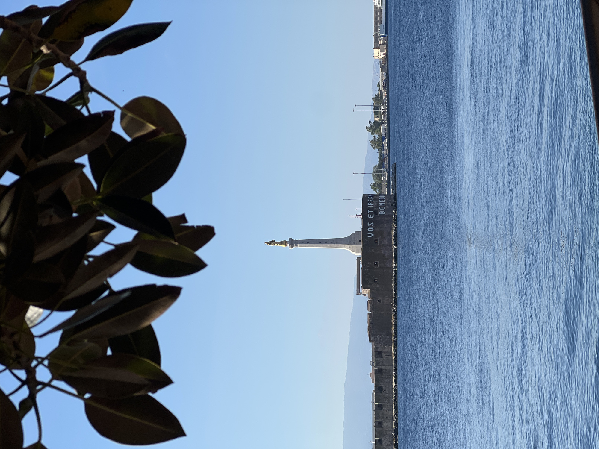 The image depicts a panoramic view of the Strait of Messina, Italy, focusing on the entrance to Messina's harbor. In the midground, the monumental column of the Madonna della Lettera is prominently featured, situated atop the historic Fort San Salvatore. The column is slender, tapering upwards, and constructed of light-colored stone or concrete. It is surmounted by a golden statue of the Madonna della Lettera, facing outwards towards the sea.
Below the column, the formidable, dark grey stone or concrete fortified wall of Fort San Salvatore stretches horizontally across the scene. Large white letters are painted on a section of this wall, clearly spelling "VOS ET IPSAM BENEDICIMUS." To the right of the column and wall, various port structures, including industrial buildings, cranes, light poles, and a white vessel (possibly a ferry) docked alongside, are visible on the distant coastline. Beyond these, a range of mountains or hills can be seen on the horizon.
The lower half of the image is dominated by the calm, deep blue waters of the Strait of Messina, exhibiting subtle ripples across its surface. The sky above is a clear, brilliant blue, indicating fair weather. The top of the image is framed by a cluster of dark, out-of-focus, broad leaves from an overhead tree branch, partially obscuring the sky. No individuals are visible in the scene.