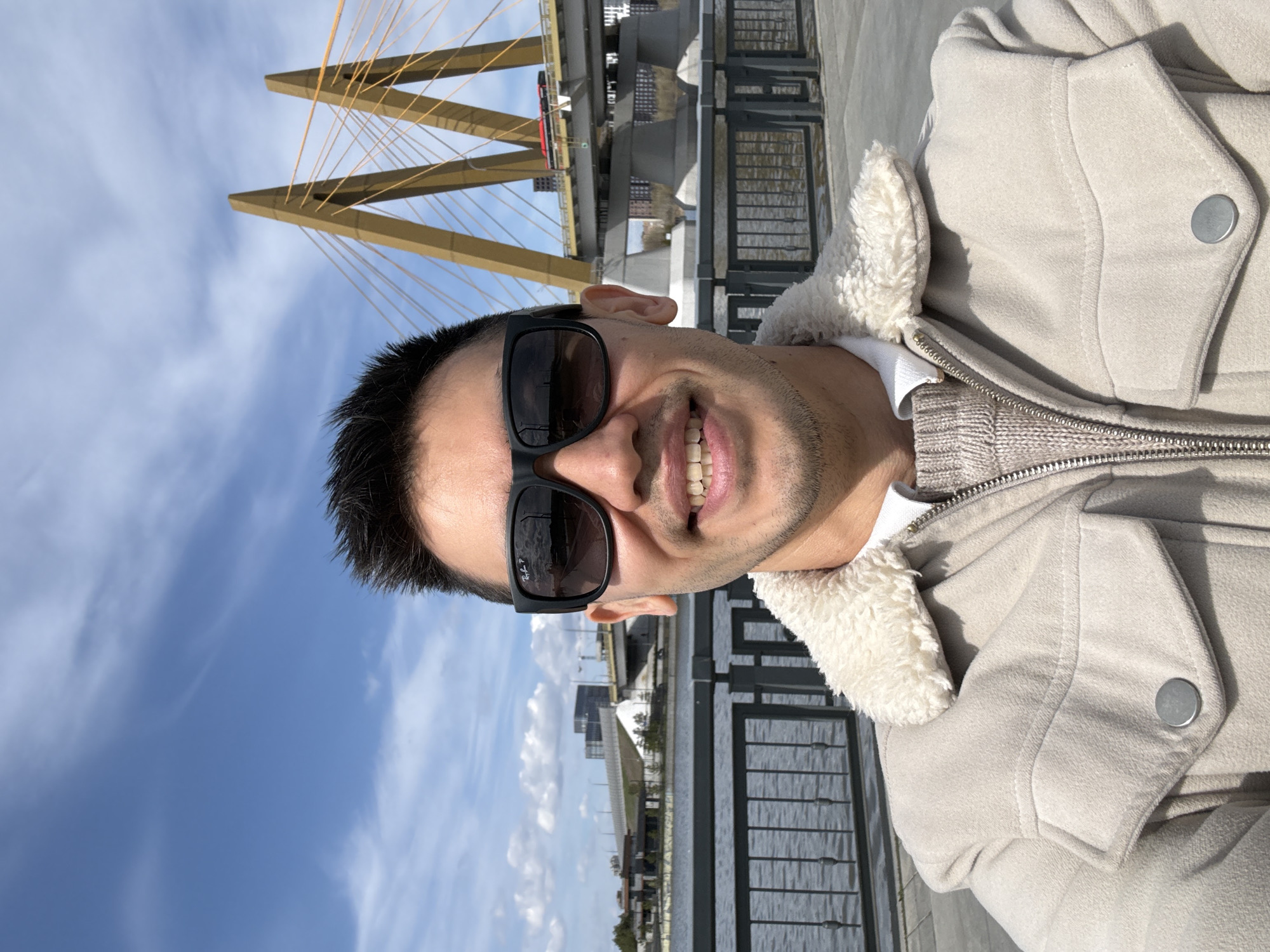 A bright, well-lit portrait-oriented selfie shows a smiling young man outdoors on a sunny day in Kazan, Russia. He is positioned in the foreground, looking directly at the camera with a wide smile, revealing his teeth. He has dark hair, dark stubble, and is wearing dark sunglasses, on which the text "Ray-Ban P" is subtly visible on the left lens. He is dressed in a light beige or off-white jacket with a prominent cream-colored shearling or faux fur collar lining, worn over a grey knitted garment, possibly a sweater or hoodie, visible around the zipper area.
The background features a clear blue sky with scattered white clouds. Behind the man, a modern cable-stayed bridge with distinctive golden-yellow, angular pylons and numerous supporting cables spans a body of water, likely the Kazanka River. The structure of the bridge is prominent, showcasing its unique design. To the right of the man, a dark railing separates the walkway from the water. In the distance, various city buildings and structures are visible under the bright sunlight. The overall scene suggests a pleasant outdoor experience, possibly along a river promenade, with the man appearing happy and relaxed.