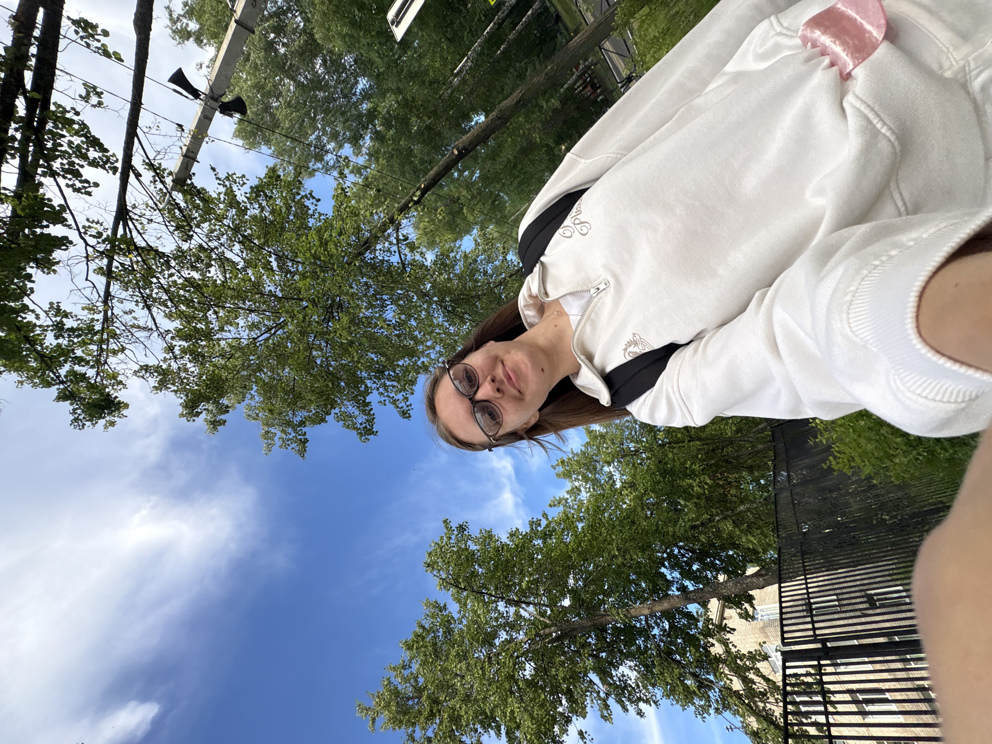 A young woman with glasses and long brown hair takes a selfie outdoors. She is wearing a white zip-up sweatshirt over a white t-shirt and a black backpack. Behind her, there are tall green trees and a bright blue sky with some wispy clouds. A black metal fence and a building with windows are visible in the background. A concrete pole with two speakers mounted on it is also present. The image appears to have been taken in the city of Chaikovsky, Russia.