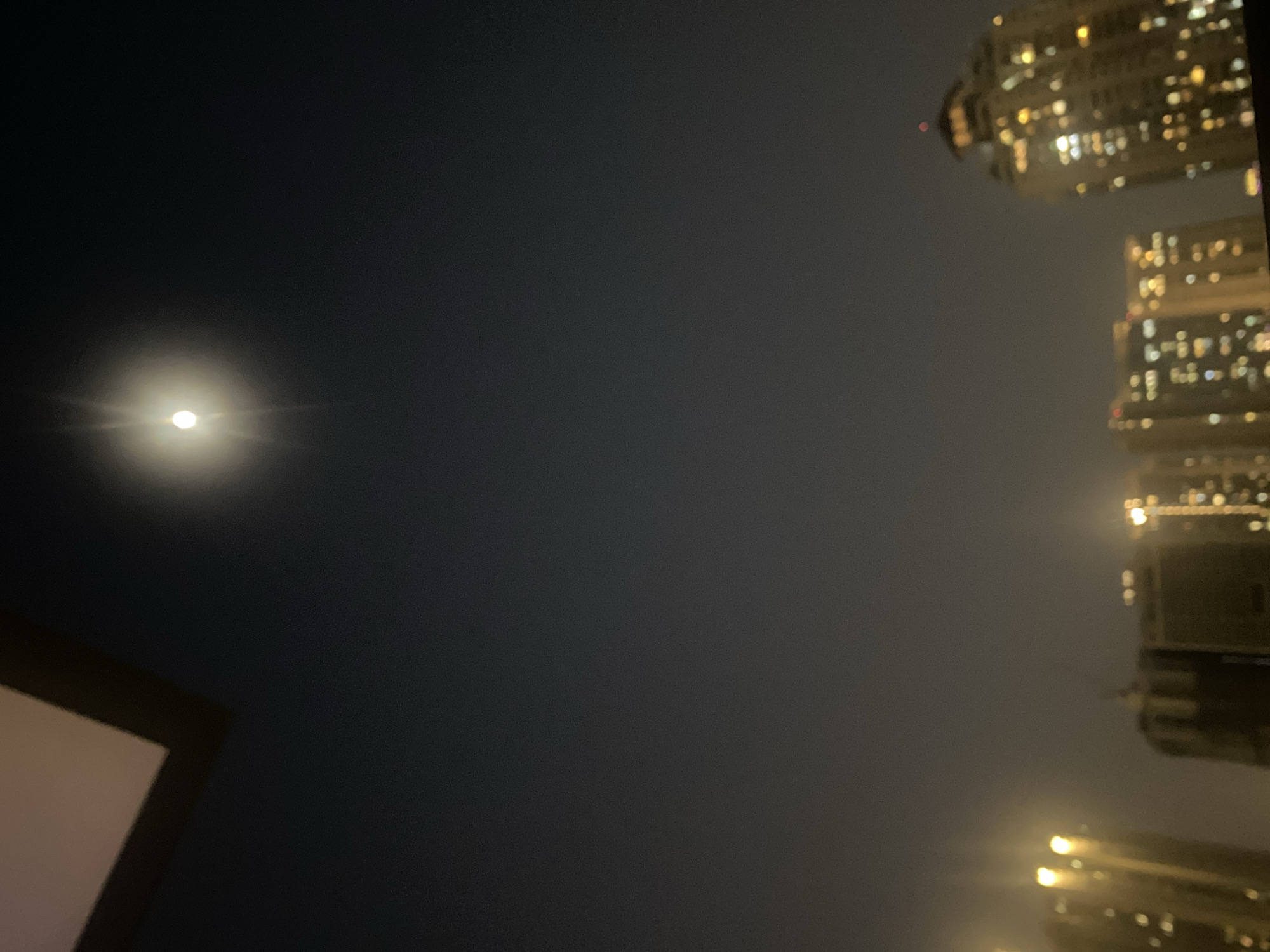 The image shows a night sky over a city. A bright, circular object, likely the moon, is visible in the upper portion of the sky, emitting a strong glow. Below the sky, illuminated buildings suggest an urban environment. Some of these buildings have a significant number of lights on, indicating they are inhabited or operational. The lower part of the image is slightly blurred, and the details of the buildings are not sharply defined. The overall impression is of a nighttime city view, possibly from an elevated position overlooking the cityscape. There are no people or specific actions depicted in the image. The presence of illuminated high-rise buildings is consistent with the landscape of Kuala Lumpur, Malaysia, known for its modern architecture and vibrant city lights.