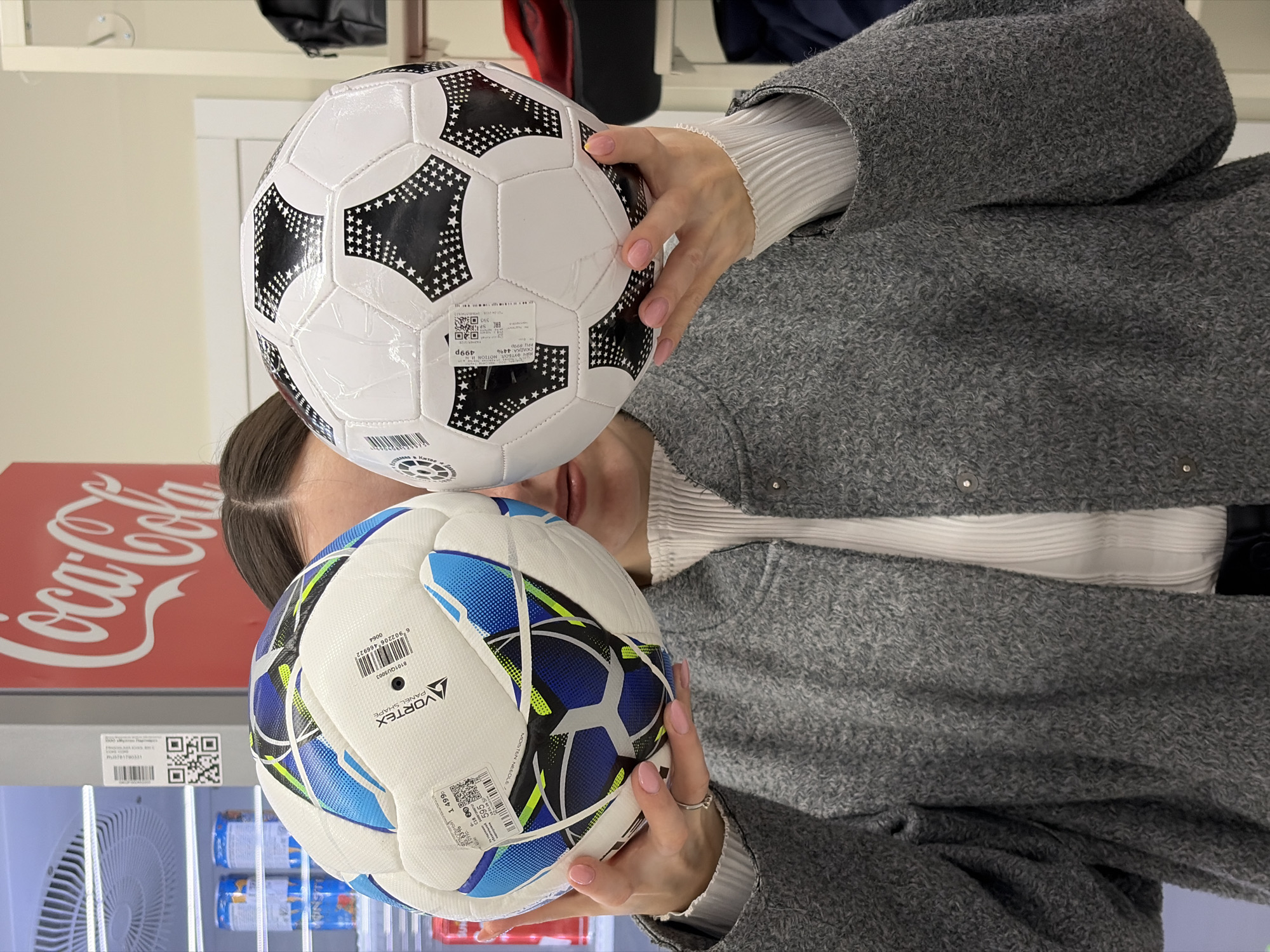 A woman in a gray wool coat stands indoors holding two soccer balls in front of her face. The top soccer ball is predominantly white with black star-shaped patterns. The bottom soccer ball is white with blue and black patterns. In the background is a Coca-Cola-branded refrigerator unit stocked with canned goods. The location is Izhevsk, Russia. The OCR text identifies a “Vortex Panel Shape” soccer ball and a price of 499 Russian rubles.