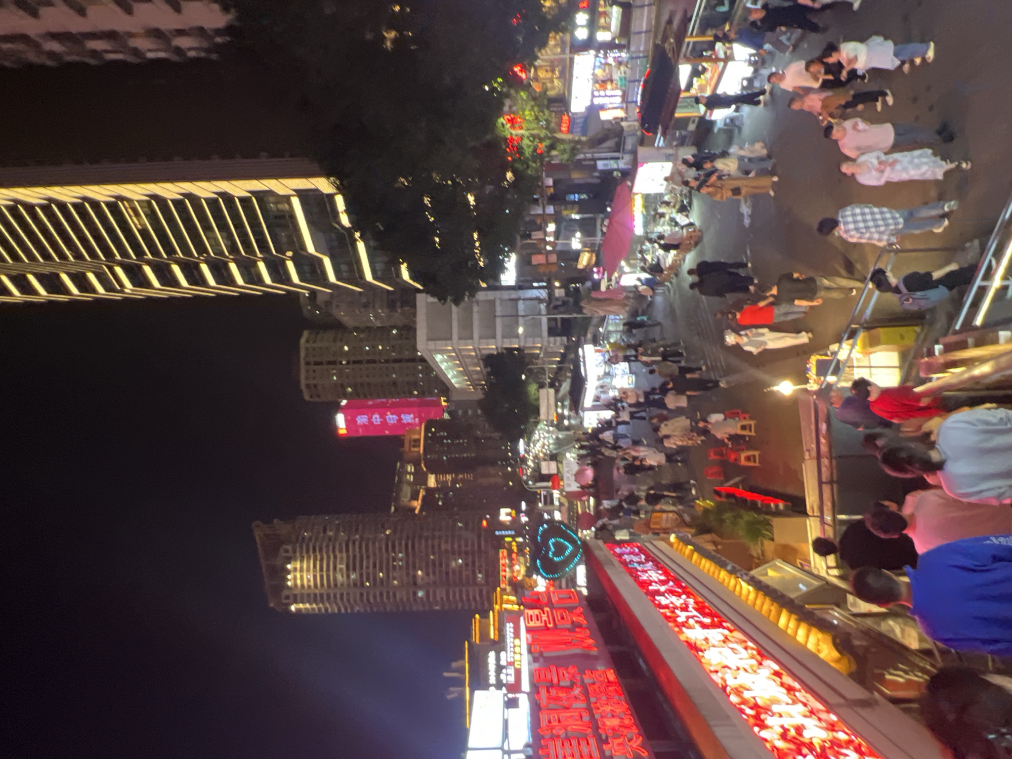 The image depicts a nighttime street scene in Yuzhong District, Chongqing, China. A dense crowd of people walks along a pedestrian street lined with brightly lit storefronts and signage in Chinese characters. Buildings of various heights are visible, including a high-rise on the left with illuminated vertical architectural details. Trees are present, partially obscuring the upper stories of the buildings. Several vendors with umbrellas are among the crowd. The scene conveys a bustling urban atmosphere with a mix of commercial and residential elements.