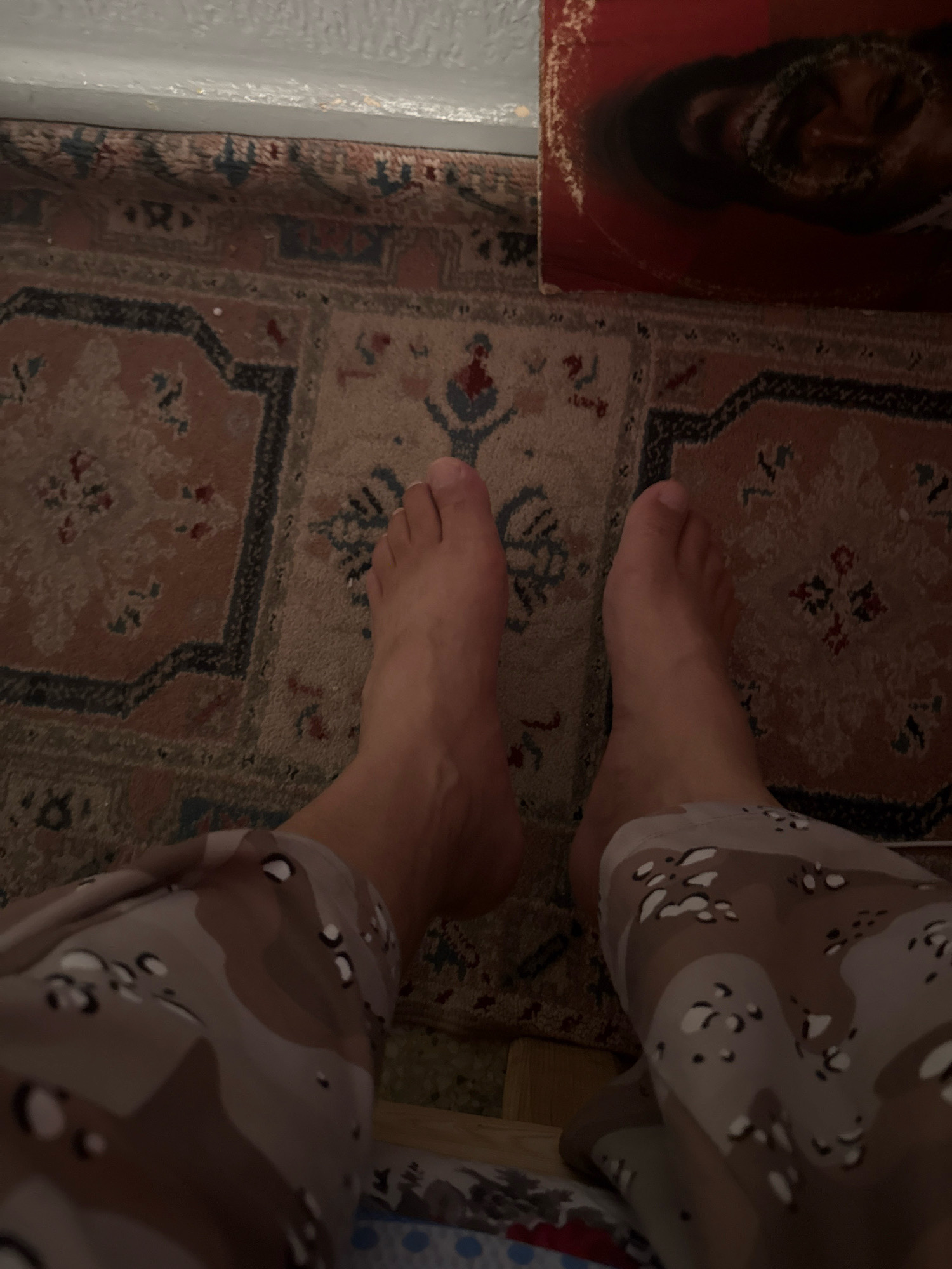 A low-angle, close-up shot shows a person's bare feet and lower legs from a seated or reclined perspective. The feet are resting on a patterned rug with an ornate, traditional Moroccan design, featuring intricate geometric and floral motifs in shades of red, brown, and blue. The person is wearing camouflage-patterned trousers on their legs. To the upper right of the frame, a portion of a vinyl record with a prominent red background and a faded image of a face is visible, suggesting the scene takes place in a residential setting. The lighting is dim, creating soft shadows. The rug and the visible portion of the vinyl record are consistent with decorative items found in homes in Fez, Morocco.