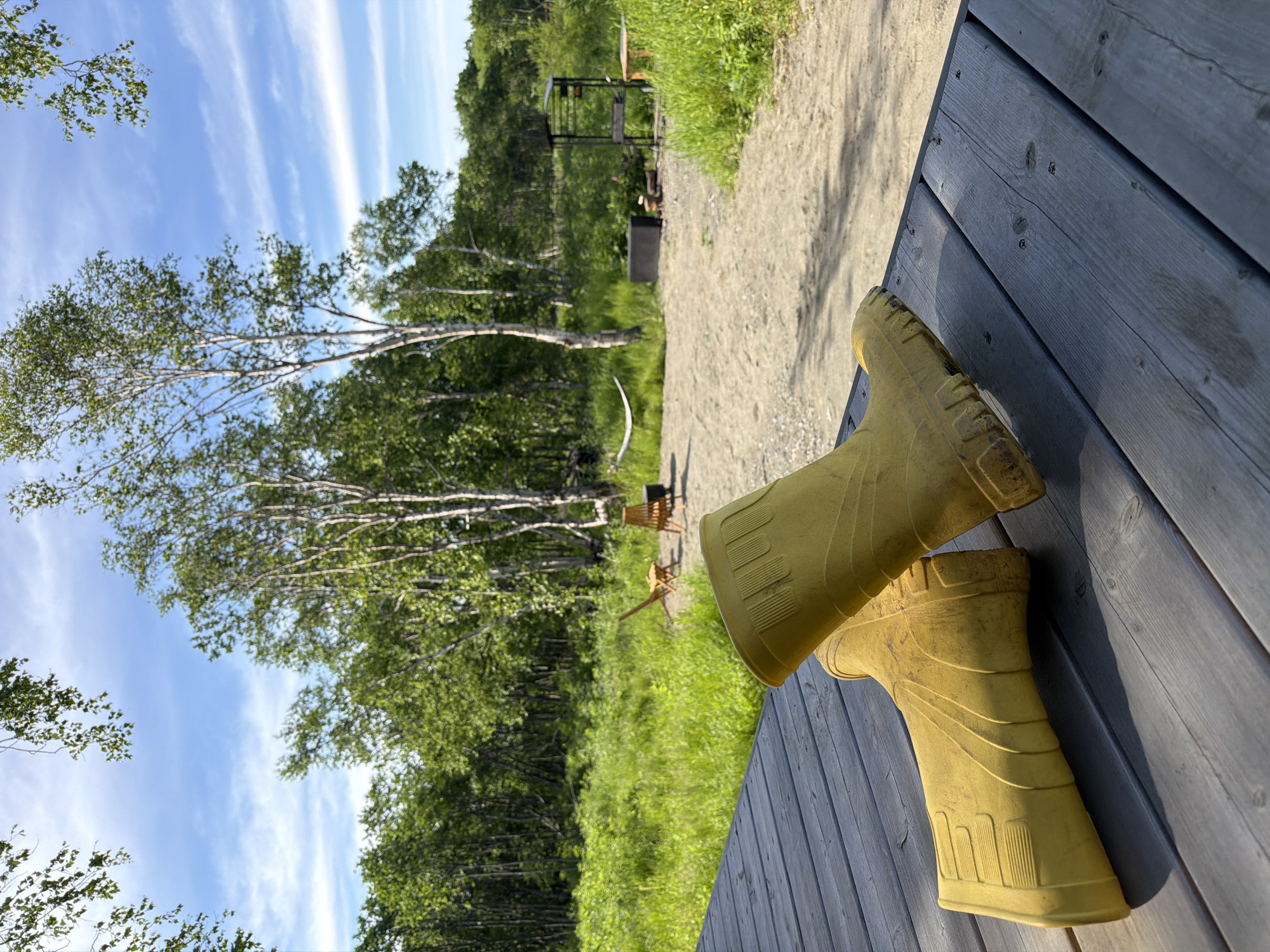 The image depicts a pair of yellow rubber boots resting on a weathered wooden deck in the foreground. Behind the deck, a dirt path leads to a grassy area with trees in the background. There are two empty wooden chairs and a black fire pit visible in the middle ground. A hammock is strung between two trees further in the distance, and a metal grill and a wooden table are situated near the edge of the tree line. The sky is blue with scattered white clouds. The scene appears to be an outdoor recreational area or campsite.