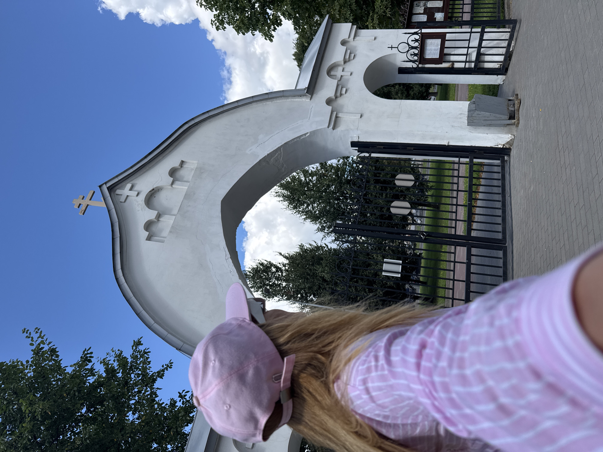 The image captures a young person wearing a pink baseball cap and a pink and white striped shirt, seen from behind, looking towards a white arched gateway with a cross on top. The gateway appears to be an entrance to a cemetery, indicated by the black metal fence and what looks like memorial plaques visible beyond the arch. The setting is outdoors with a clear blue sky dotted with white clouds, and lush green trees surrounding the area. The ground is paved with grey interlocking bricks. The overall scene suggests a bright, sunny day. The image does not contain any information to pinpoint the location to Saint Petersburg, Russia, or any specific city.