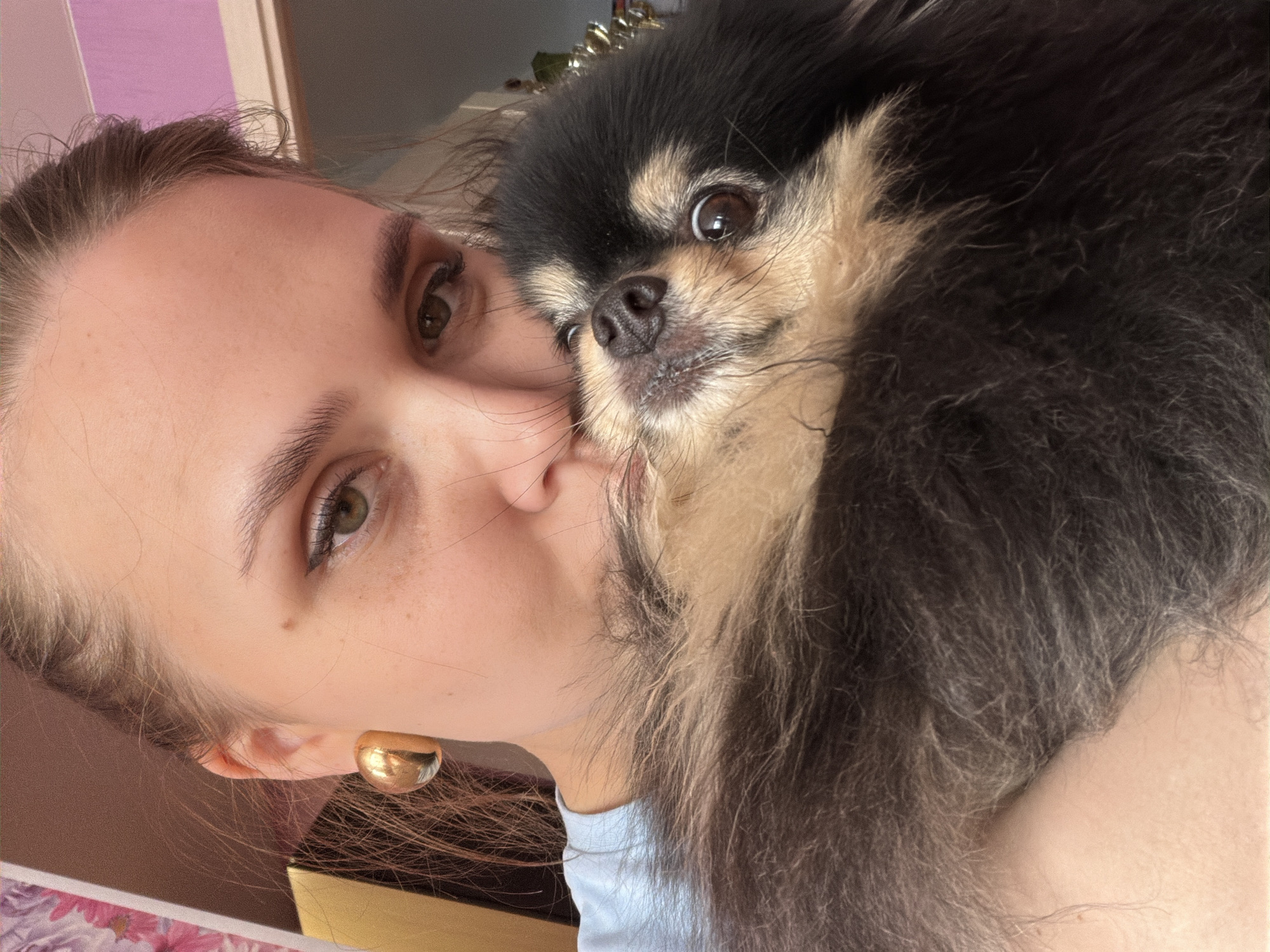 A close-up vertical image depicts an adult human female and a small canine, photographed in an indoor domestic setting within Chaikovsky, Russia. The female, displaying light skin, brown hair pulled back, hazel eyes, and a visible gold-colored half-hoop earring on her left ear, wears a light blue garment. She holds a small, long-haired dog, identified as a black and tan Pomeranian-type breed, against her face, with the dog's head resting near her chin. Both subjects face forward, looking towards the camera. The background is out of focus, showing sections of a warm-toned wall, a floral pattern, and a bright pink wall portion on the upper right.