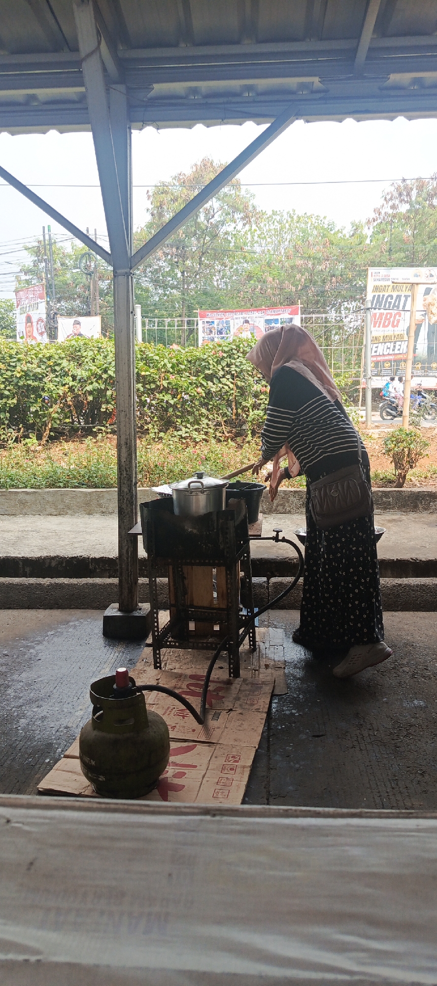 A woman wearing a hijab and a striped shirt is cooking outdoors in Sukaresmi, Indonesia. She is standing next to a metal stove with a pot on it. A gas cylinder is positioned on a piece of cardboard on the ground. Behind her, there are bushes and trees, and in the distance, banners and a motorcycle are visible. The scene appears to be under a covered area with metal supports.