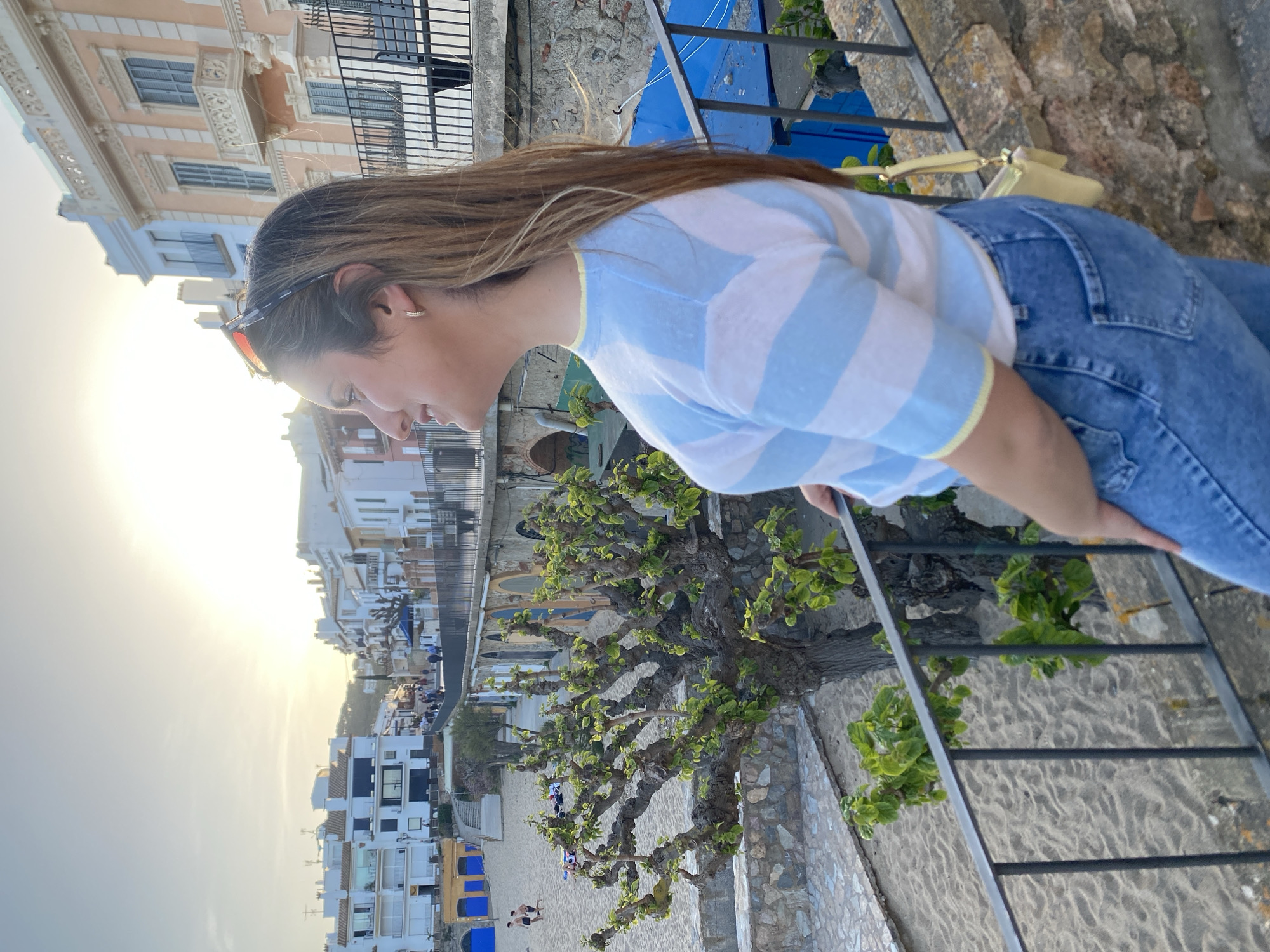 A photograph shows a woman in a striped blue and white shirt and jeans leaning over a black iron railing, looking down towards a beach and town. She has long brown hair and is wearing sunglasses on her head. The beach has light-colored sand, and several people are visible on it. Blue beach umbrellas are also present. The town is composed of white buildings with a mix of architectural styles. The photo appears to be taken during the late afternoon, as the sun is low in the sky, creating a bright glow. In the foreground, between the woman and the beach, is a tree with green leaves. A stone wall and steps lead down towards the beach. In the upper background, a building with ornate architectural details is visible. The photograph's location is Calella De Palafrugell, Spain.