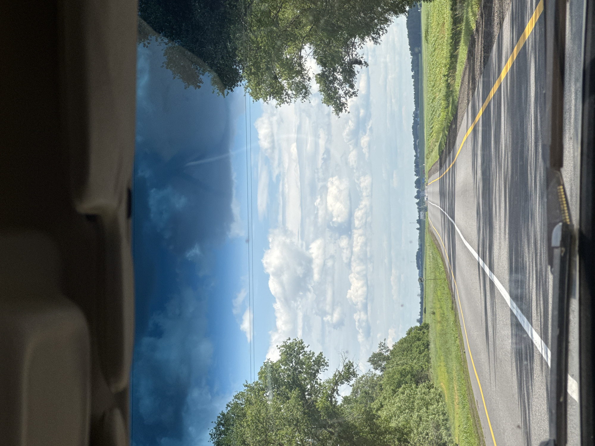 A point-of-view shot from inside a vehicle shows a highway stretching into the distance under a partially cloudy sky. The road is paved with asphalt and features a white center line and a yellow line on the right edge. Lush green trees line both sides of the road, with fields of crops visible beyond them. The sky is a mix of blue and white clouds, with some darker clouds suggesting a potential for rain. The scene appears to be set in a rural area, likely within the city of Oktyabrsky Sel'skiy Sovet, Belarus, given the landscape and road infrastructure. There are no people or animals visible in the image.