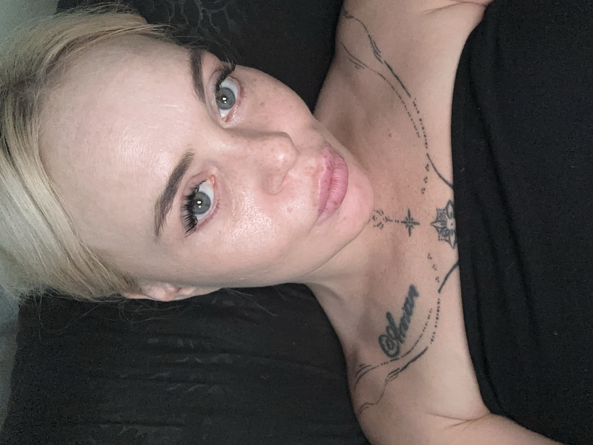 A close-up, overhead selfie of a woman with blonde hair pulled back and light blue eyes. Her skin is fair with some freckles and minor blemishes. She is wearing a strapless black garment. She has tattoos on her chest and collarbone area, one of which appears to be the word "Amor" in cursive on her upper chest. The background is dark and appears to be a soft surface, possibly fabric or leather. The lighting is dim and likely artificial. The image was taken indoors.