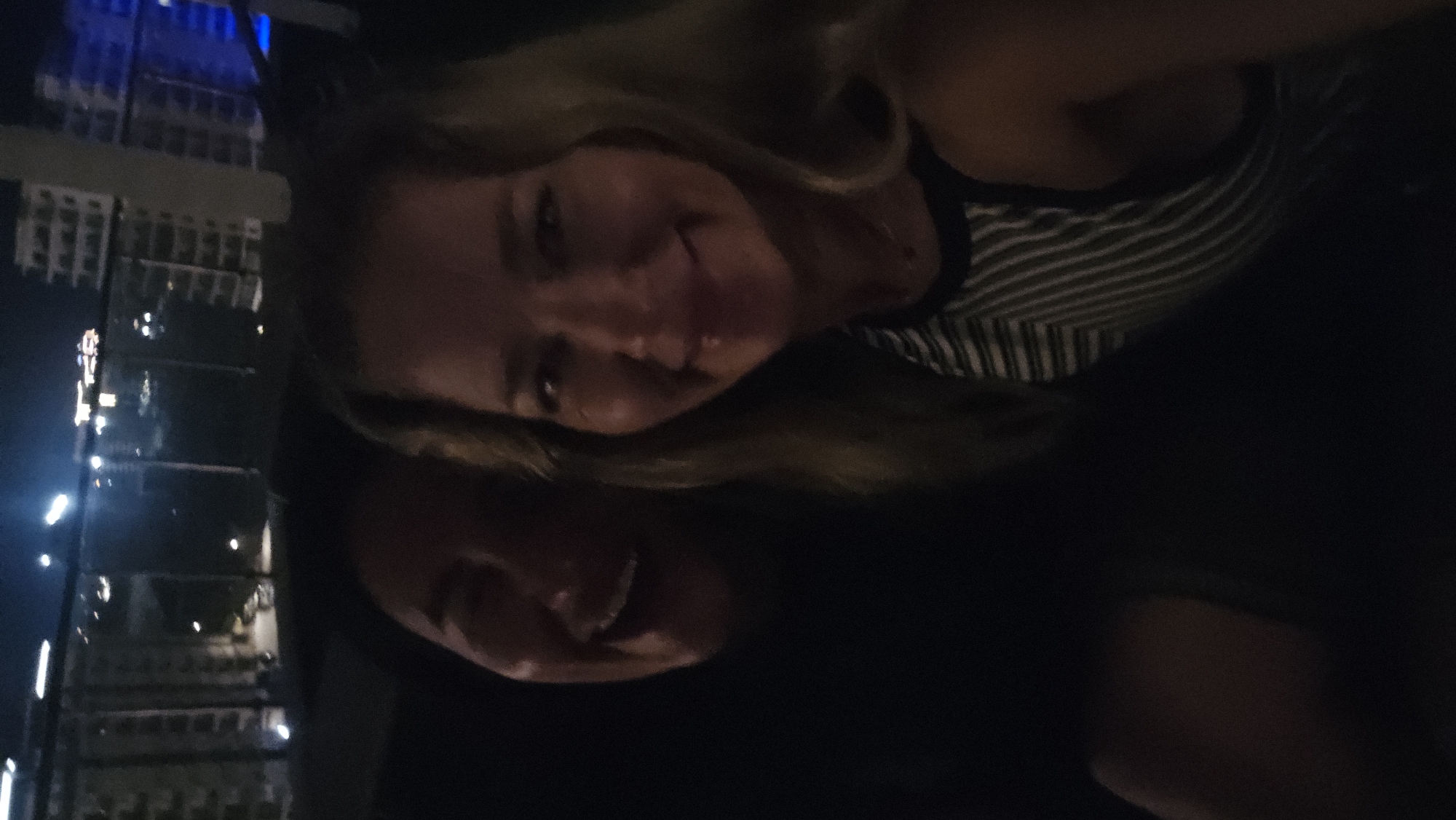Two individuals, a man and a woman, are captured in a close-up selfie taken at night. The man is on the left, with his face partially obscured by darkness and his mouth open in a wide laugh. The woman is on the right, smiling and looking towards the camera. She is wearing a striped tank top. Behind them, a blurred cityscape is visible with illuminated buildings and lights, suggesting they are on a balcony or rooftop overlooking the city. The overall impression is a candid and joyful moment shared between two people. The location is the city of Unknown, Unknown.