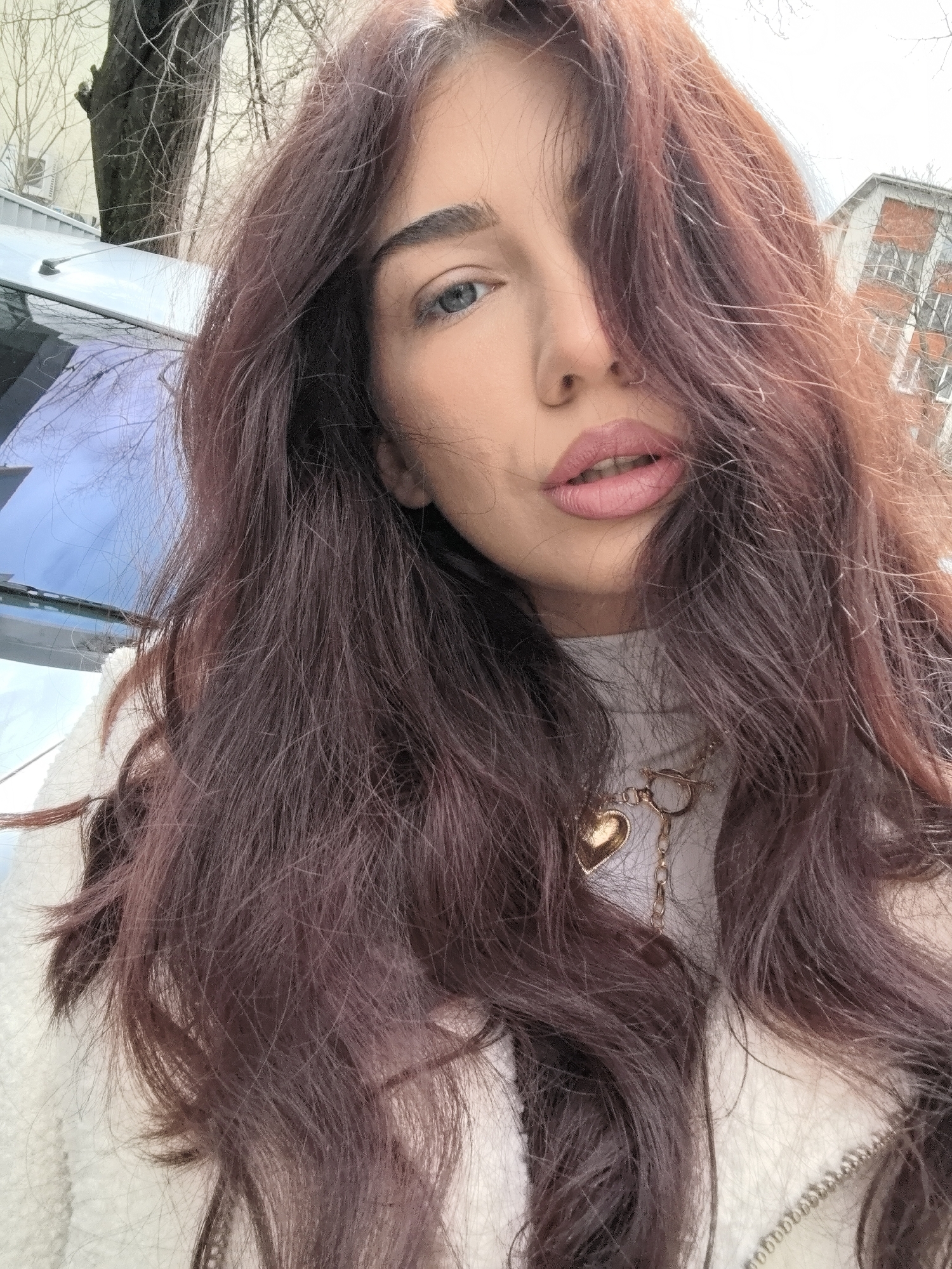 The image presents a close-up portrait of a single female subject in the city of Stara Zagora, Bulgaria. The subject has long, wavy, dark reddish-brown hair, part of which falls across the right side of her face. Her blue eyes are directed towards the camera, and her lips are painted with pink lipstick, slightly parted to reveal the upper teeth. She wears a light-colored turtleneck sweater, and a gold-toned necklace with a heart-shaped pendant and a toggle clasp is visible around her neck. The background features urban elements, including a partial view of a light-colored vehicle's roof and windshield to the left, bare tree branches and trunks, and segments of buildings: a lighter-colored structure on the far left and a brick-faced building with windows on the right. The sky overhead appears bright.
