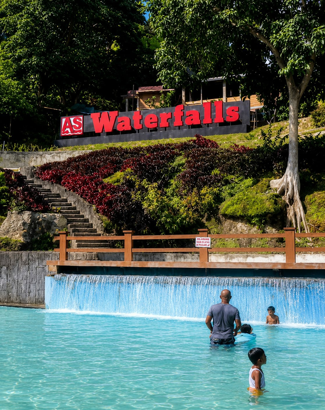 In Silaiyajae, Indonesia, a man and three children are seen in a bright turquoise swimming pool. The pool features a man-made waterfall cascading into the water from a wooden structure. Behind the pool, a set of stone stairs leads up a hill covered in lush green foliage and vibrant red plants. A large sign with red lettering that reads "AS Waterfalls" is prominently displayed on the hillside. The sky above is a clear blue, indicating a sunny day.