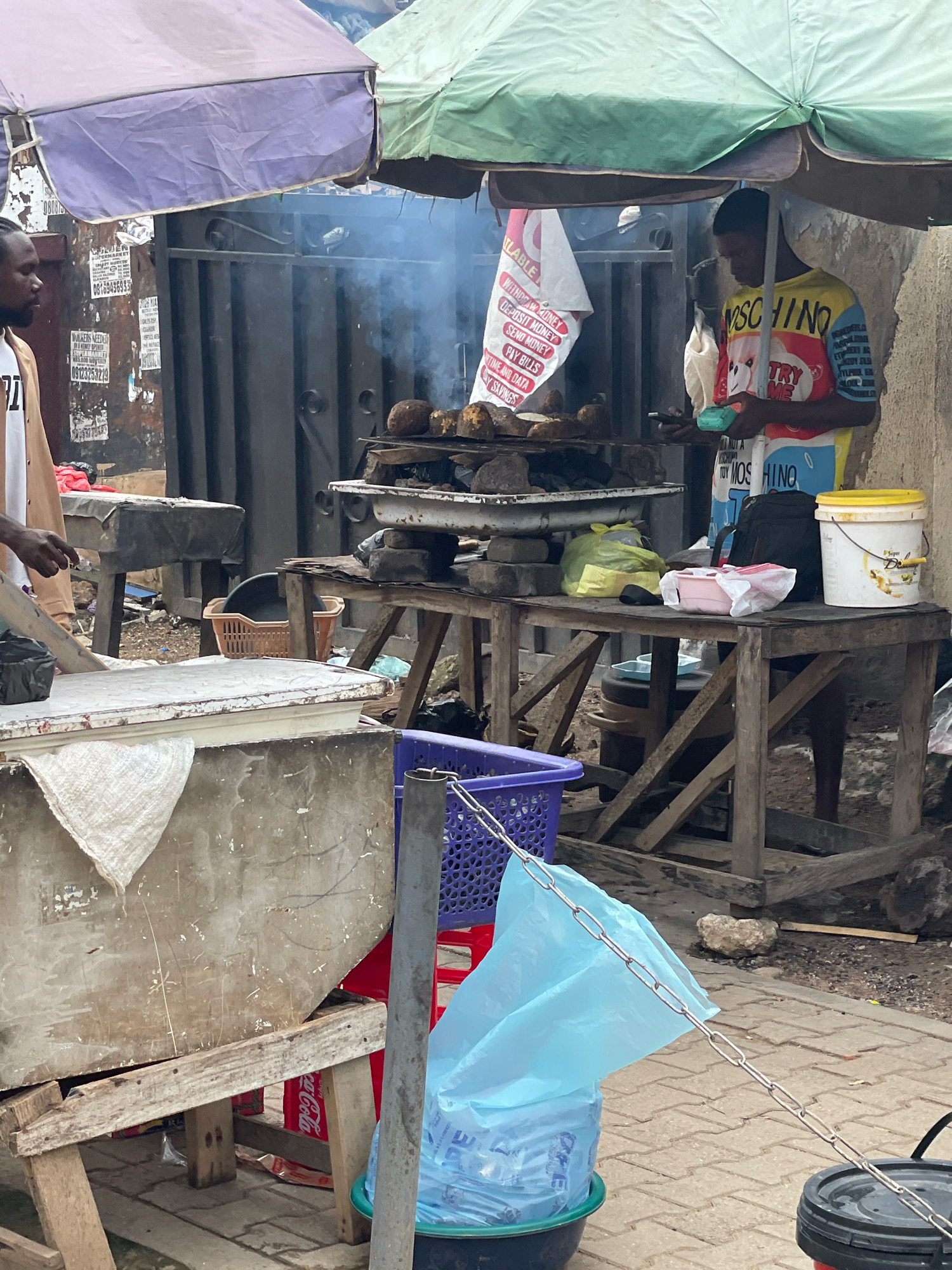 In a bustling outdoor market in Abuja, Nigeria, a young man stands under a makeshift canopy formed by a green and purple umbrella, tending to a charcoal grill. Smoke billows from the grill, where several round food items, possibly yams or plantains, are being roasted. The vendor, wearing a colorful, graphic-print t-shirt, is engaged with a customer or an associate, holding a mobile phone and a small, green electronic device.
Adjacent to the grill, a wooden table is laden with various items, including a white bucket with a yellow lid, a pink food container, and a yellow plastic bag. In the foreground, a large, weathered cooler or storage unit occupies the left side of the frame, draped with a white cloth. To the right of the cooler, a purple plastic basket sits on the ground. The overall scene depicts a typical street food vendor setup, with goods and cooking apparatus arranged for efficient service. The background shows a metal gate and posters affixed to a wall, suggesting a dense urban environment.