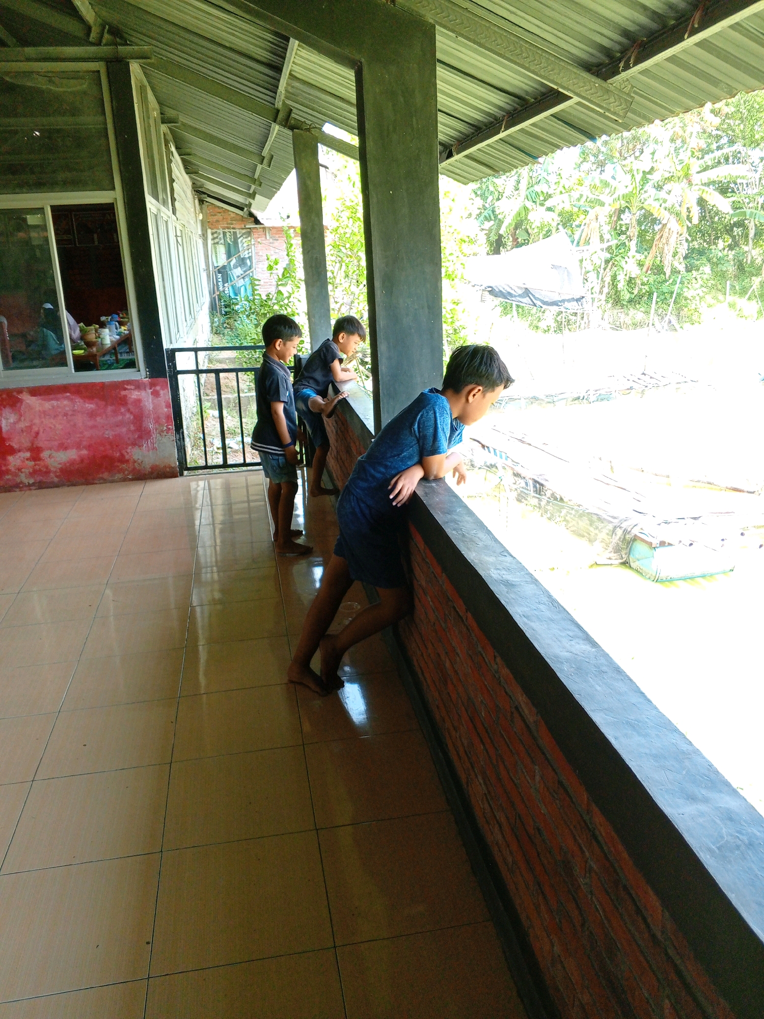Three young boys are standing along a railing, looking out over a body of water. The boy on the left, wearing a black t-shirt and blue shorts, is looking down and to the right. The middle boy, in a black t-shirt and blue shorts, is leaning on the railing and looking to the right. The boy on the right, wearing a blue t-shirt and dark shorts, is leaning on the railing with his arms crossed and looking to the right. The setting appears to be outdoors, with a tin roof overhead and lush greenery visible in the background. The floor is tiled. The overall scene suggests a casual outing, possibly at a place with a view or an animal enclosure. This image was taken in Kayen, Indonesia.