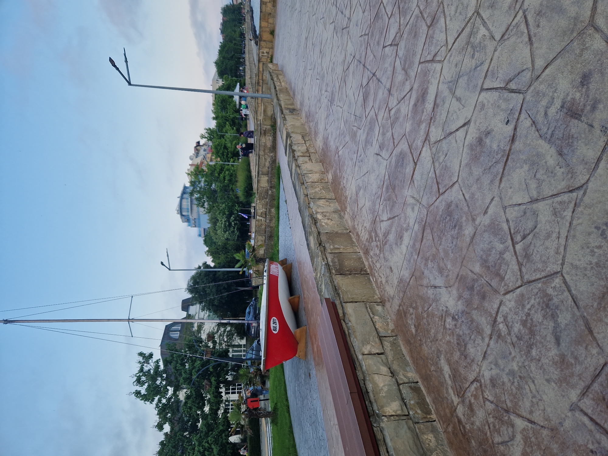 The image displays an outdoor scene, likely a promenade or park area adjacent to a body of water, under an overcast sky. In the foreground, a textured paved pathway, characterized by irregular flagstone-like patterns in grey and brown tones, occupies the lower and right sections. A low stone wall, constructed from roughly cut, light brown/grey blocks, runs along the left edge of the pathway, topped by a dark brown wooden bench seat.
In the midground, a red and white sailboat, devoid of sails but equipped with a mast and rigging, is exhibited on wooden stands on a grassy patch to the left of the path. The hull displays the text "BUL 2611" in white and "ПРИБОЙ" (Priboy, likely in Cyrillic) in white on the red section. Behind the boat, green trees and smaller palm-like plants partially obscure a white building with a dark roof. Several individuals are present in this area; one person in a blue shirt is visible walking away from the camera near the boat, and others are further in the background. A tall, grey streetlight with a dual-headed fixture rises on the right side of the paved path.
The background reveals more buildings, including one with a distinctive blue roof, positioned beyond a dense line of trees. Multiple indistinct figures are visible on an elevated pathway in the distance. The sky is a pale blue with scattered grey clouds, dominating the upper portion of the image. No specific interactions or detailed actions of the approximately 10-15 visible people are discernible; most appear to be walking or standing.