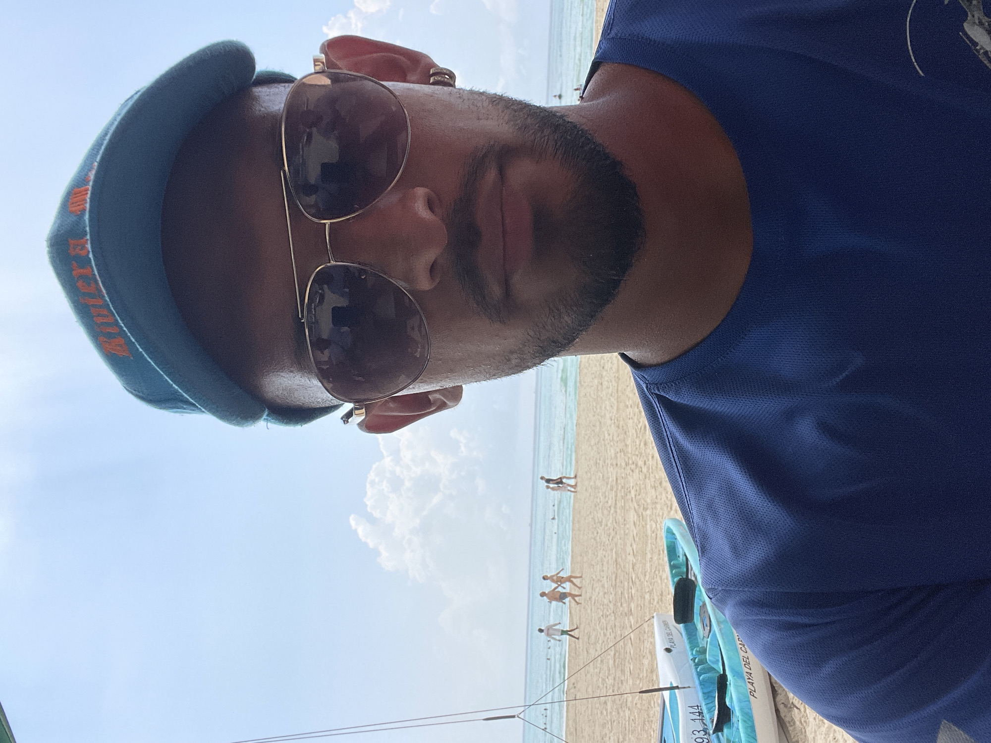 The image is a selfie taken on Playa del Carmen beach in Mexico. A man with a beard and sunglasses is wearing a blue hat and a blue shirt. Several people are walking on the sandy beach. In the background, there are clouds in the sky and blue ocean water. Part of a light blue boat is visible.