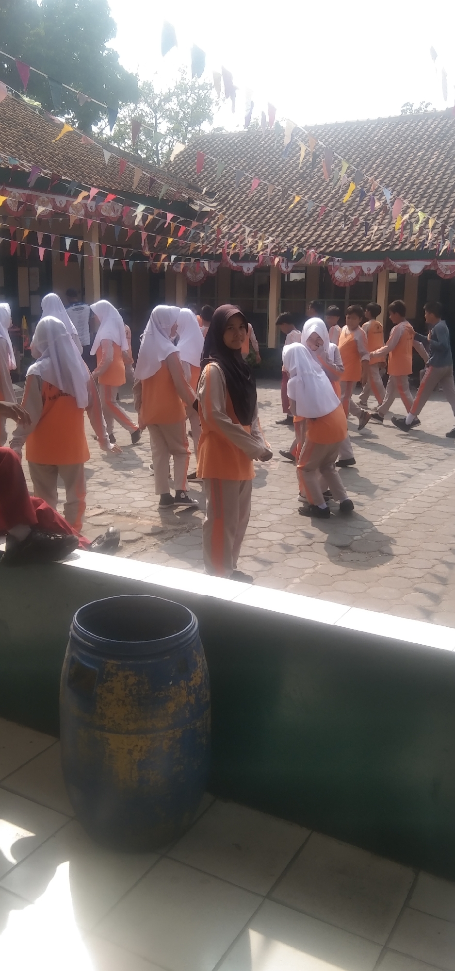 In the city of Rahayu, Indonesia, a group of schoolchildren, predominantly girls wearing orange uniforms and headscarves, are gathered in a schoolyard. The yard is paved with hexagonal tiles. In the background, a building with a tiled roof and open windows is visible, decorated with a string of colorful triangular flags. The weather appears sunny. In the foreground, a large blue and yellow barrel sits near a low green wall. The children are interacting and moving, suggesting some form of organized activity or play.