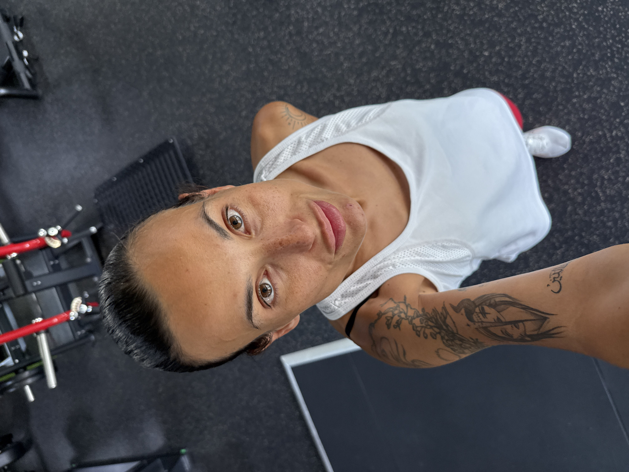 A woman is taking a selfie in a gym. She is looking up at the camera, with her hair pulled back. She is wearing a white tank top and red shorts. She has tattoos on her arm. The gym has black rubber flooring and some weight machines in the background. The image does not provide any information about the location in the city of Pleven, Bulgaria.