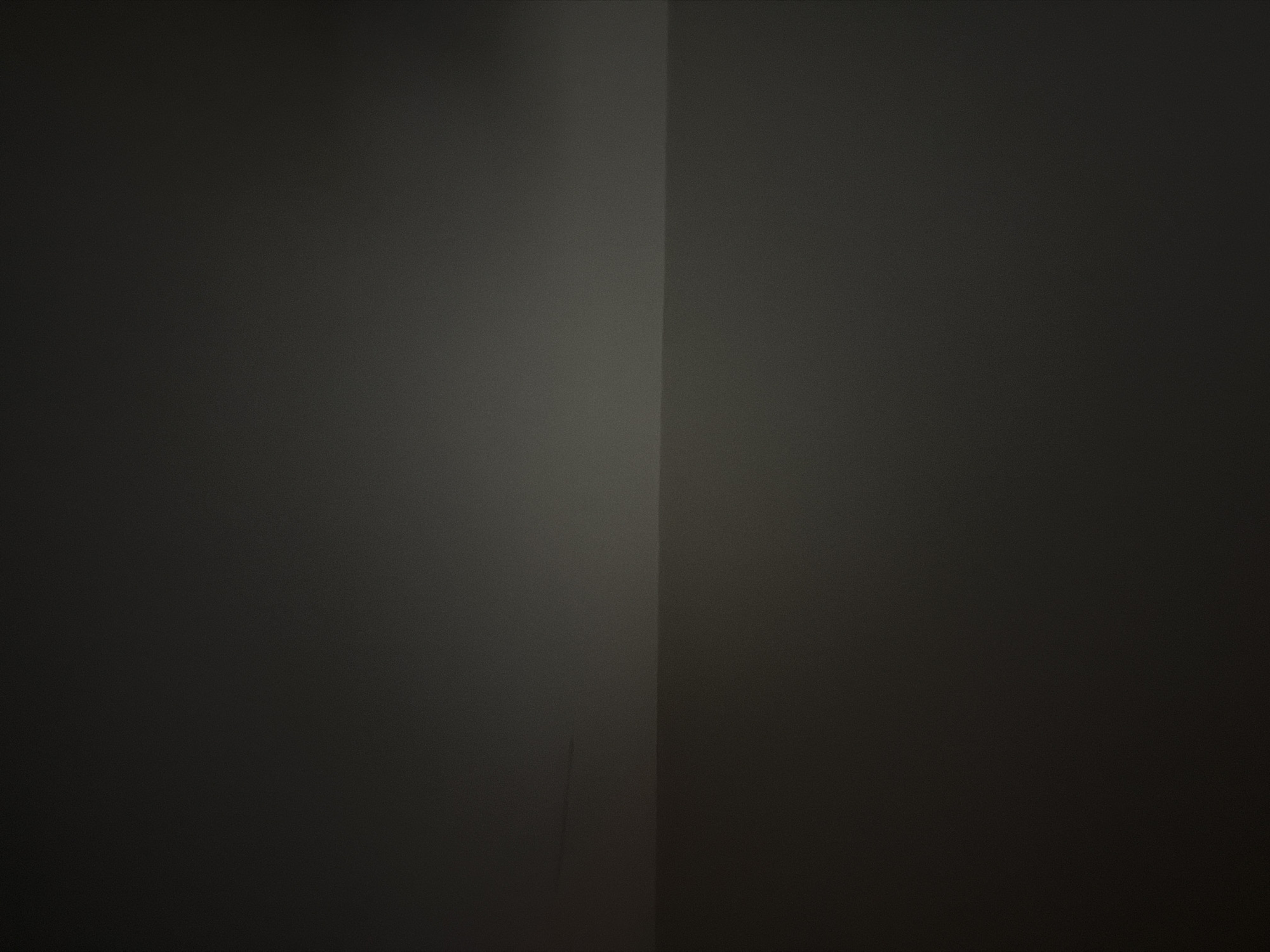 The image is an abstract, dark, and minimally lit composition, primarily featuring shades of gray and black. A distinct horizontal line divides the frame, with the upper portion appearing slightly lighter and more diffuse, potentially suggesting a subtle light source or gradient. The lower portion is darker and more uniform. There are no discernible objects, people, or actions present, and the image lacks any specific environmental cues that would indicate a location within The Hague, Netherlands, or any other city.