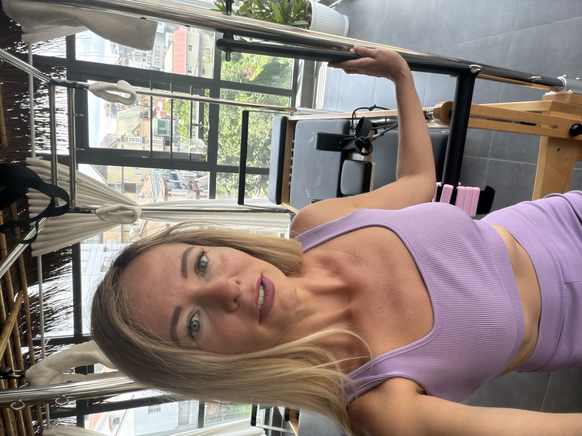 In Da Lat, Vietnam, a blonde woman with blue eyes and fair skin is captured in a selfie-style portrait. She is wearing a purple ribbed sports bra and matching bottoms. Her left arm rests on a Pilates reformer machine, its wooden frame and metal components visible. The background reveals a large window offering a view of Da Lat's urban landscape, characterized by red-tiled roofs and multi-story buildings, interspersed with lush greenery. The room's ceiling features a bamboo-like structure, and white curtains hang near the window. The lighting suggests it is daytime, with natural light illuminating the scene.