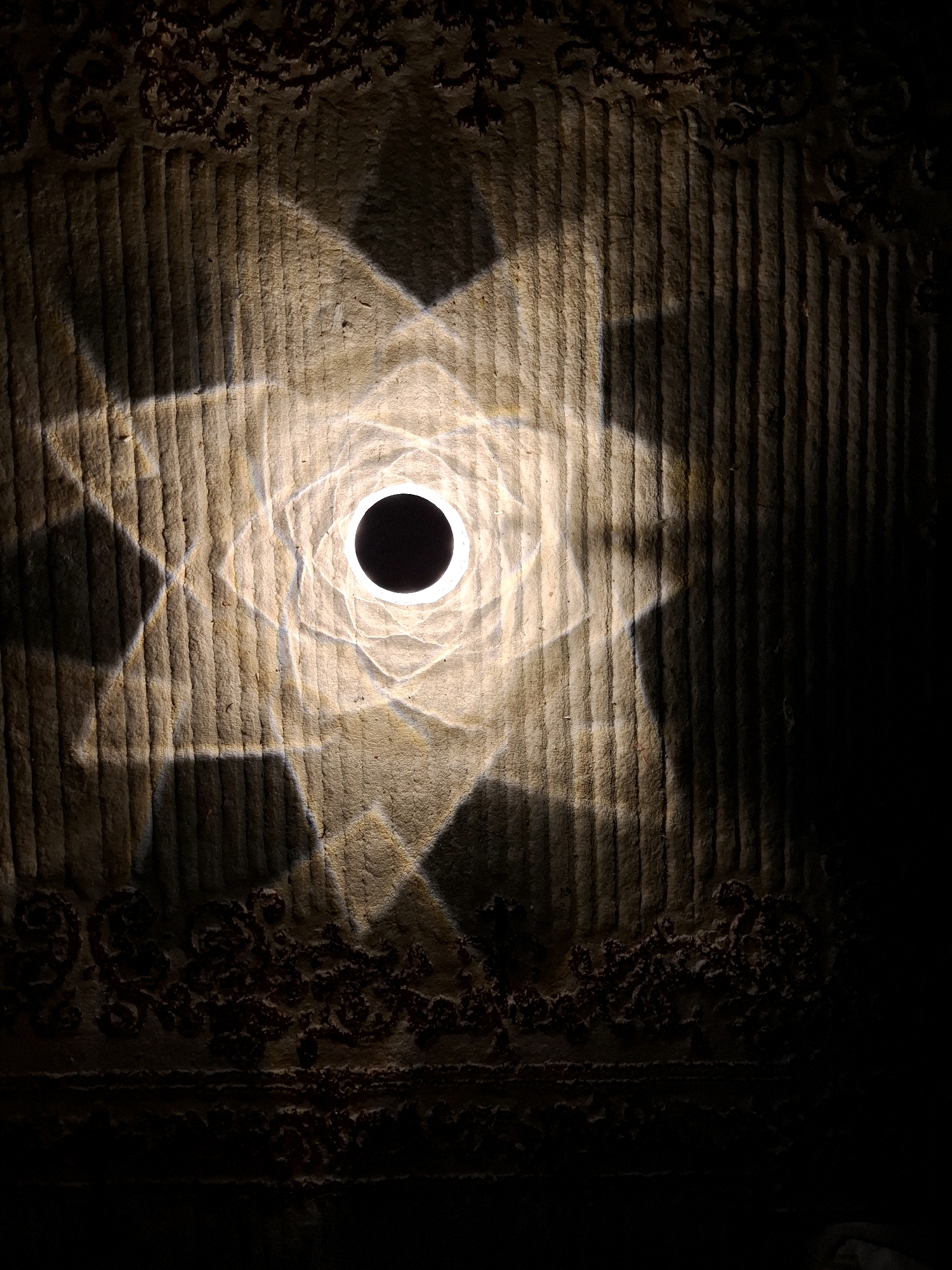 The image displays a close-up, overhead view of a textured, possibly thatched or woven, surface illuminated by a central, bright, circular light source. This light casts a complex, star-like pattern of shadows and illuminated geometric shapes across the surface. The surface itself has a rough, fibrous texture with distinct vertical ridges and is bordered at the top and bottom by a darker, decorative band with intricate, organic patterns. The overall impression is of an artistic play of light and shadow on a rustic material. There are no people or identifiable landmarks of Abu Dhabi present in the image.
