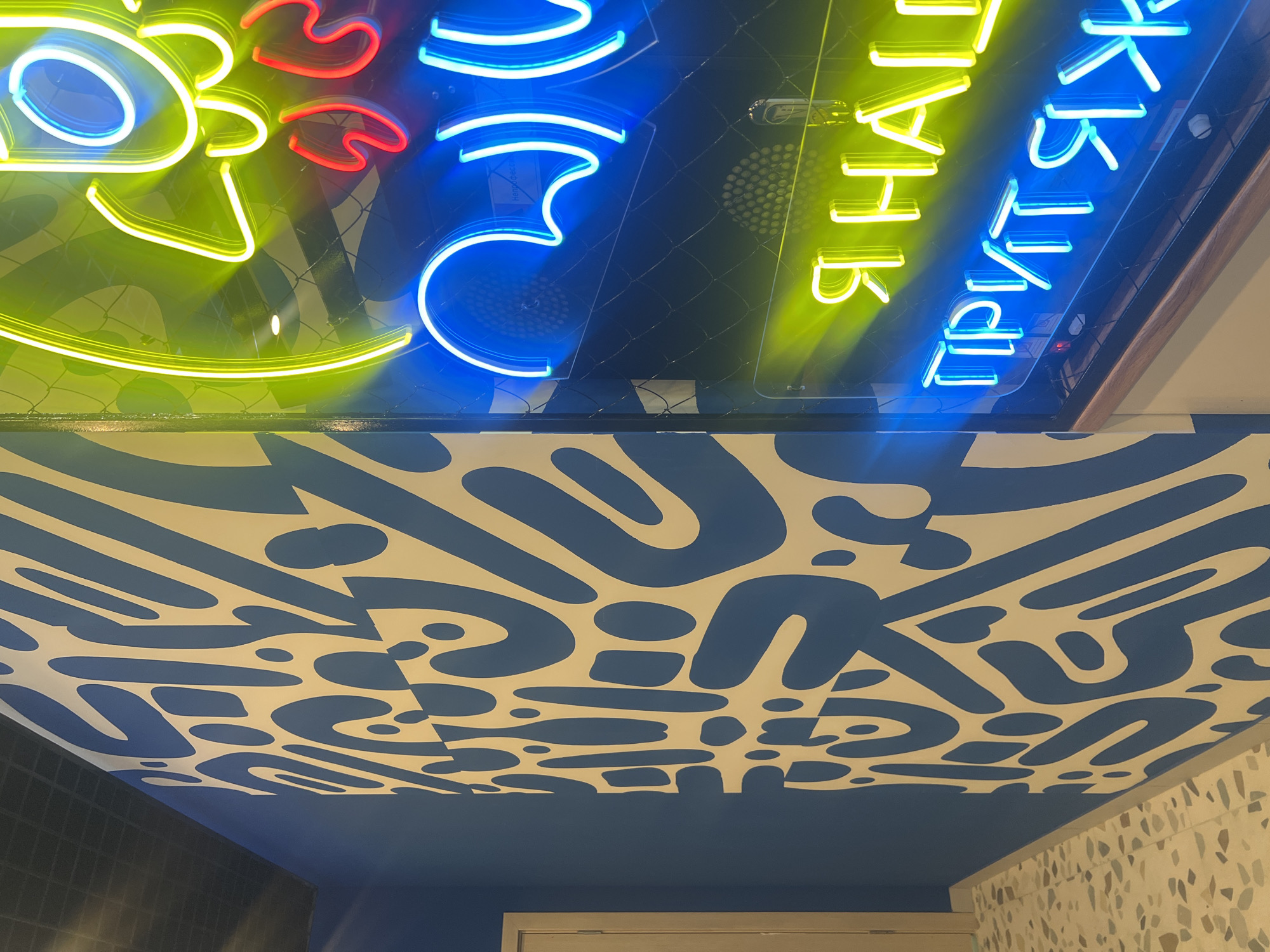 The image shows an interior view of a space in Chaikovsky, Russia. The ceiling is painted with a blue and white abstract pattern of amoeba-like shapes. Above the painted ceiling, there are neon signs. One sign has a green and red design reminiscent of a cartoon figure and the other shows blue and yellow Cyrillic characters, likely spelling out a restaurant name. The lower part of the wall is tiled.